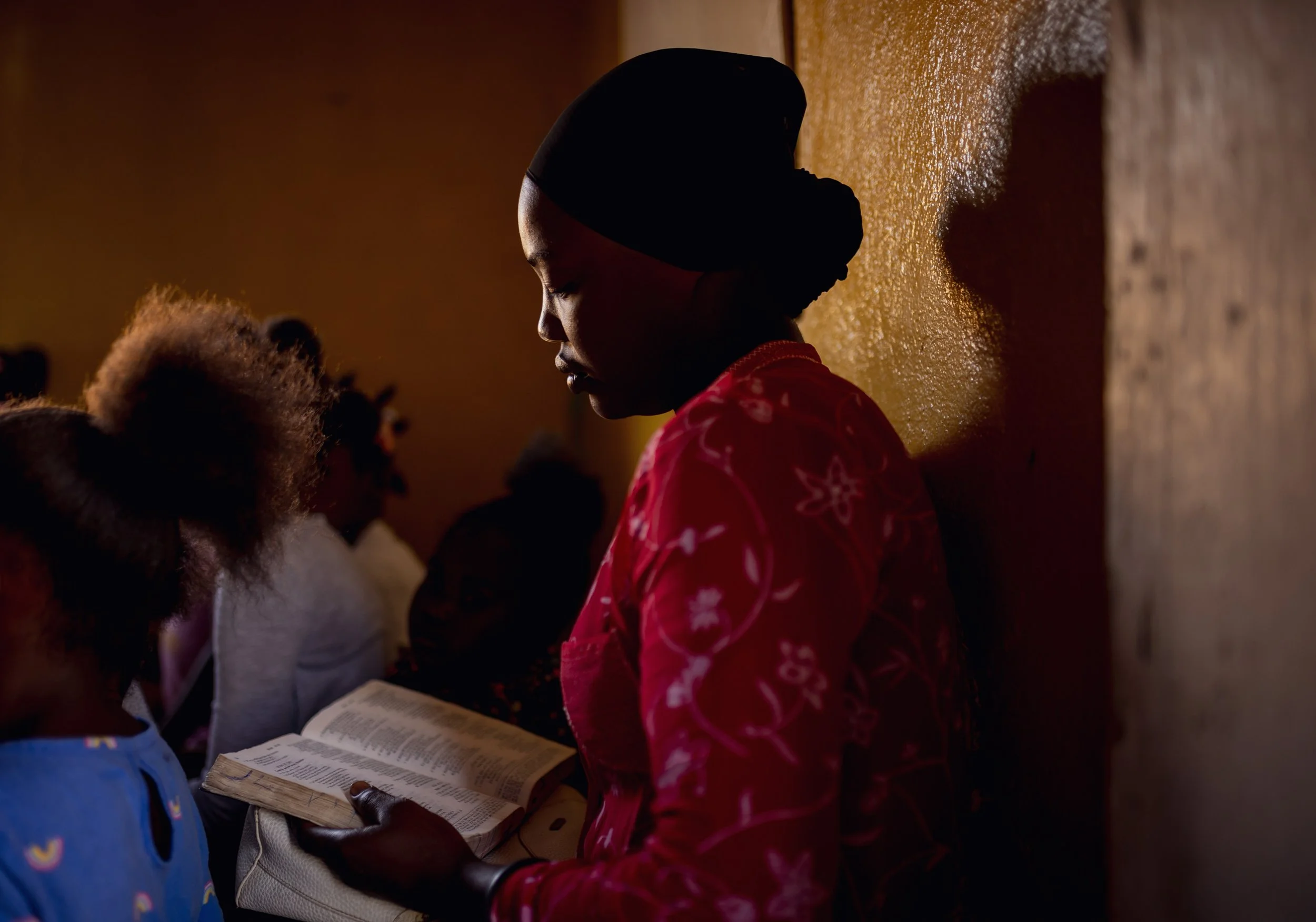 A woman reading a Bible in a church with children, with warm lighting and a textured orange wall behind her.