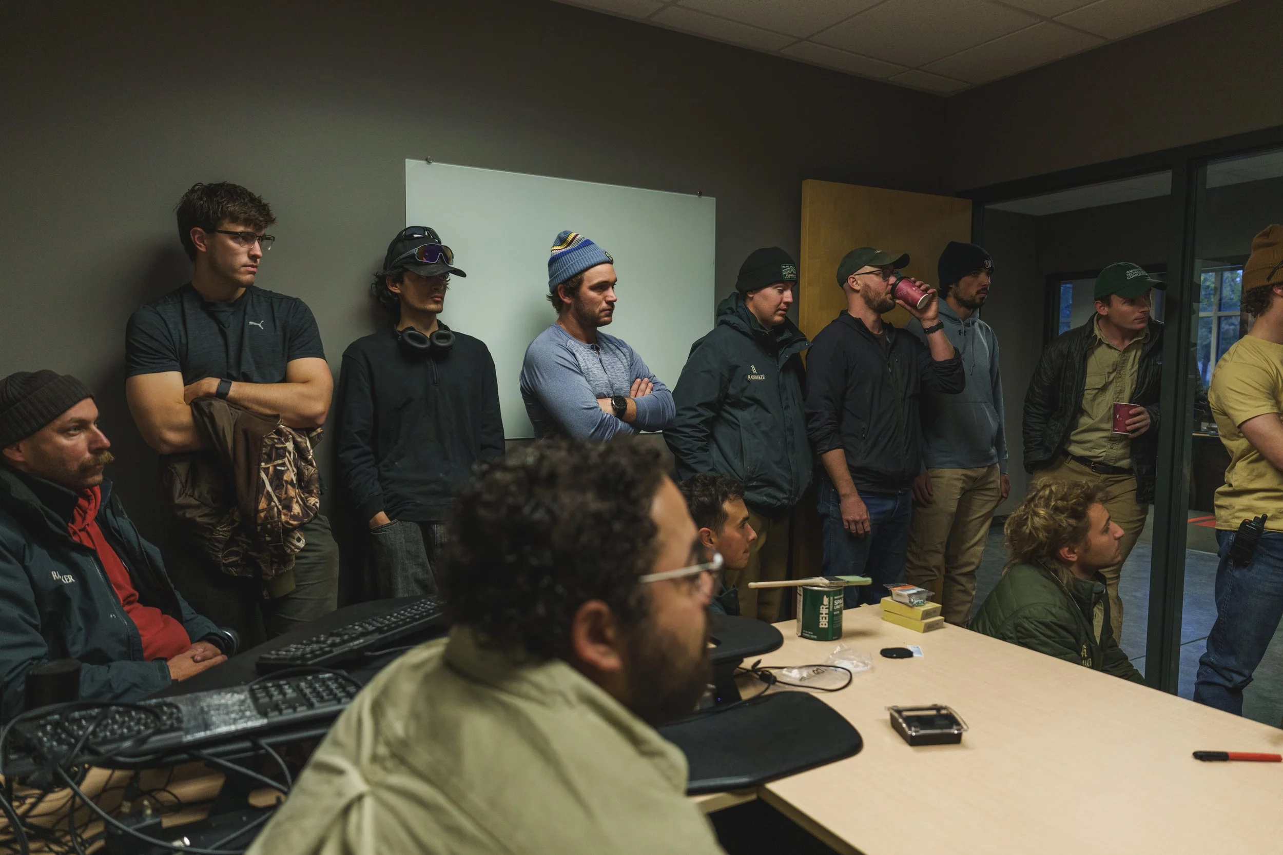A group of people gathered in a conference room, some standing along the wall and others seated at a table, listening to a presentation or discussion.