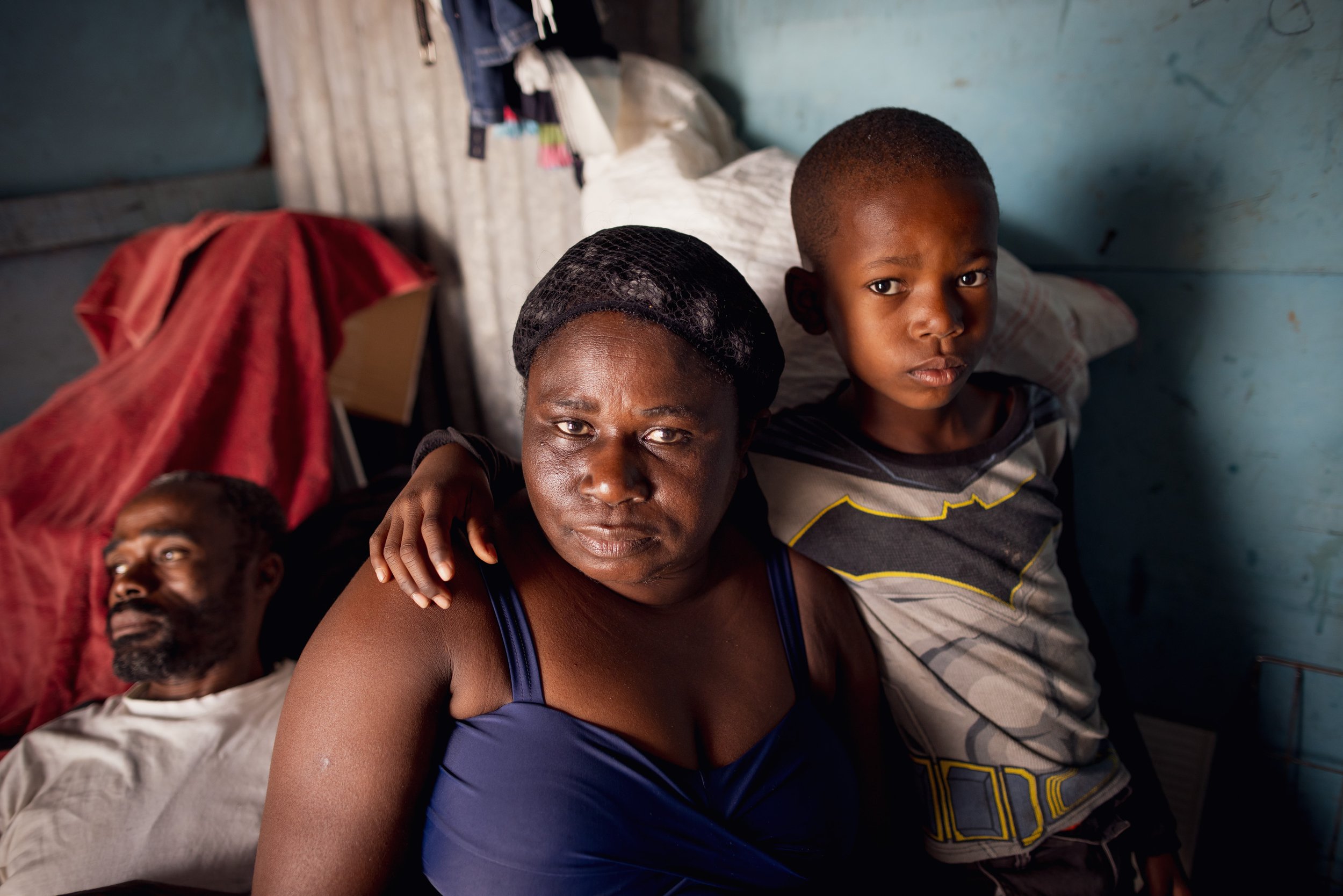 A woman and a young boy sit together inside a modest, possibly rural dwelling, with a man lying on a bed in the background. The woman has her arm around the boy, and they both look directly at the camera with serious expressions.