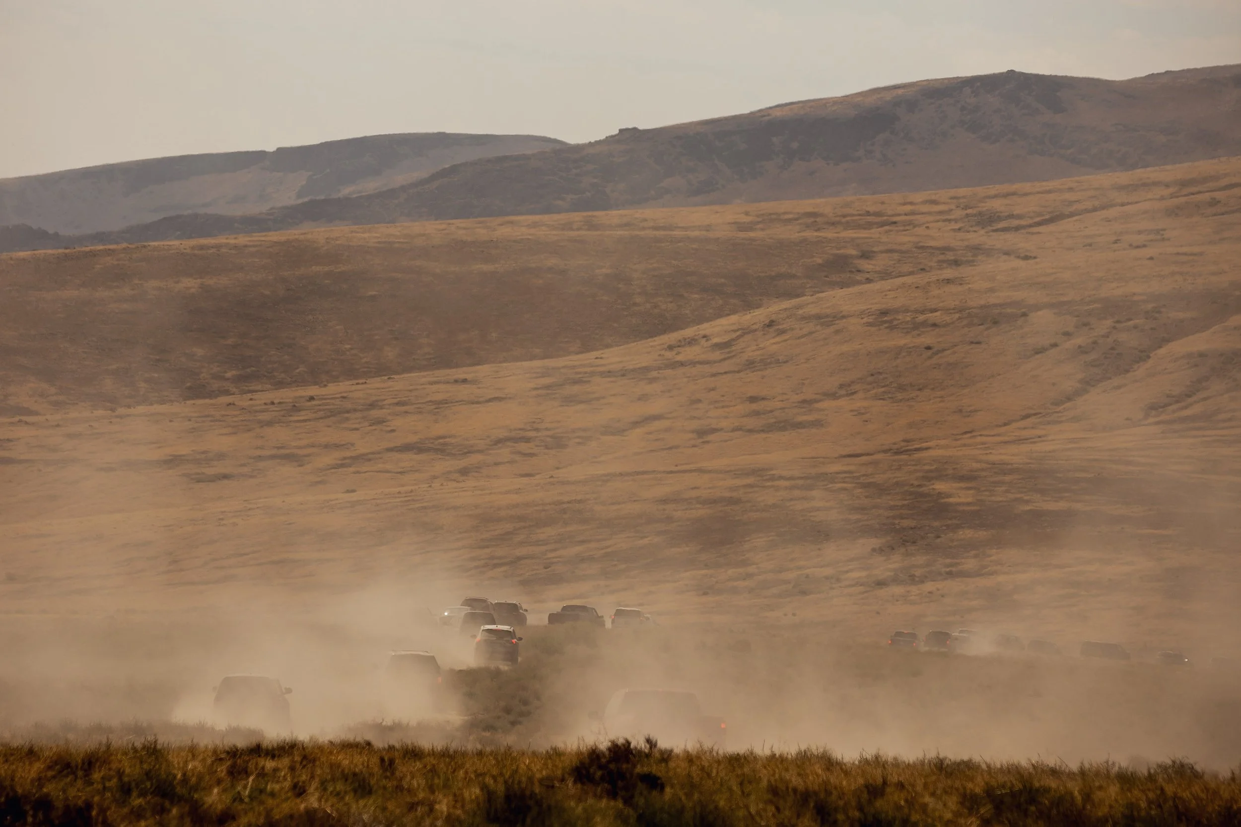A dusty road in a rural area with multiple cars driving away, surrounded by dry grassy fields and rolling hills or mountains in the distance under a partly cloudy sky.