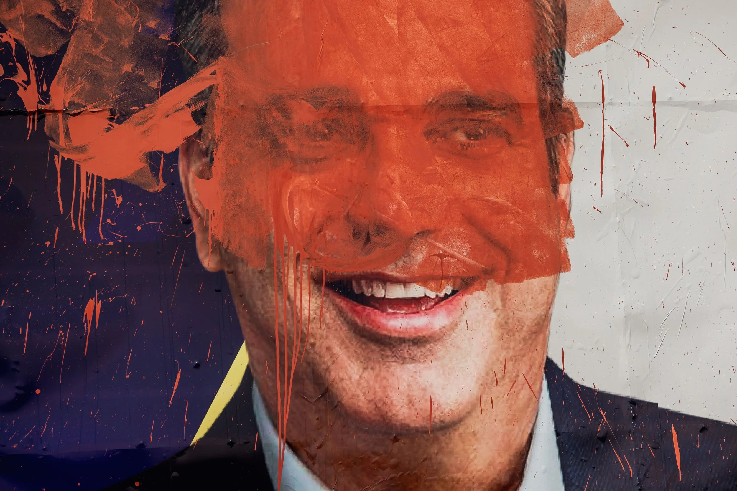 A smiling man with glasses wearing a suit, overlaid with red paint splatters and streaks, creating an abstract artistic effect.