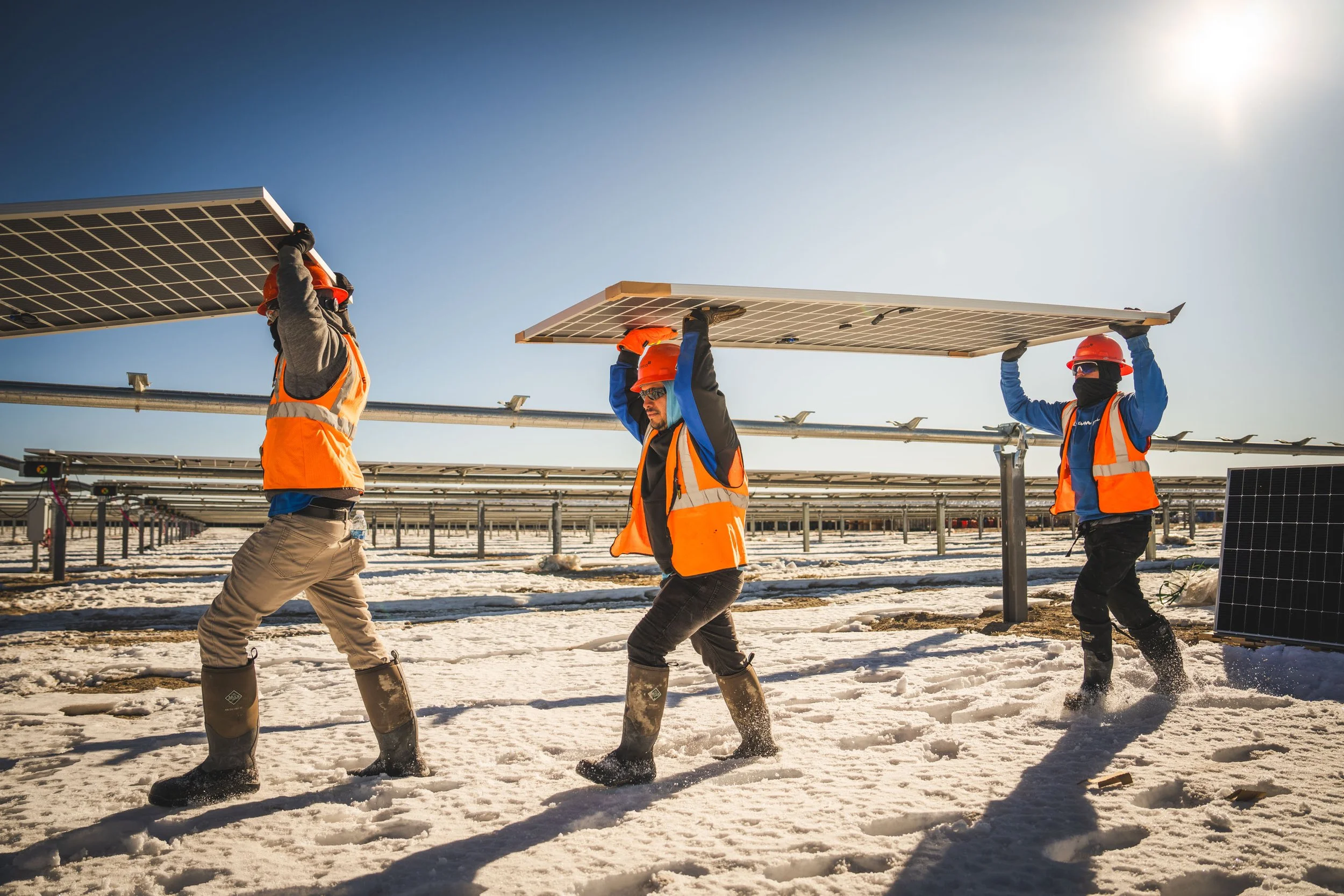 Three workers in safety gear installing solar panels on a snowy field under a clear sky.