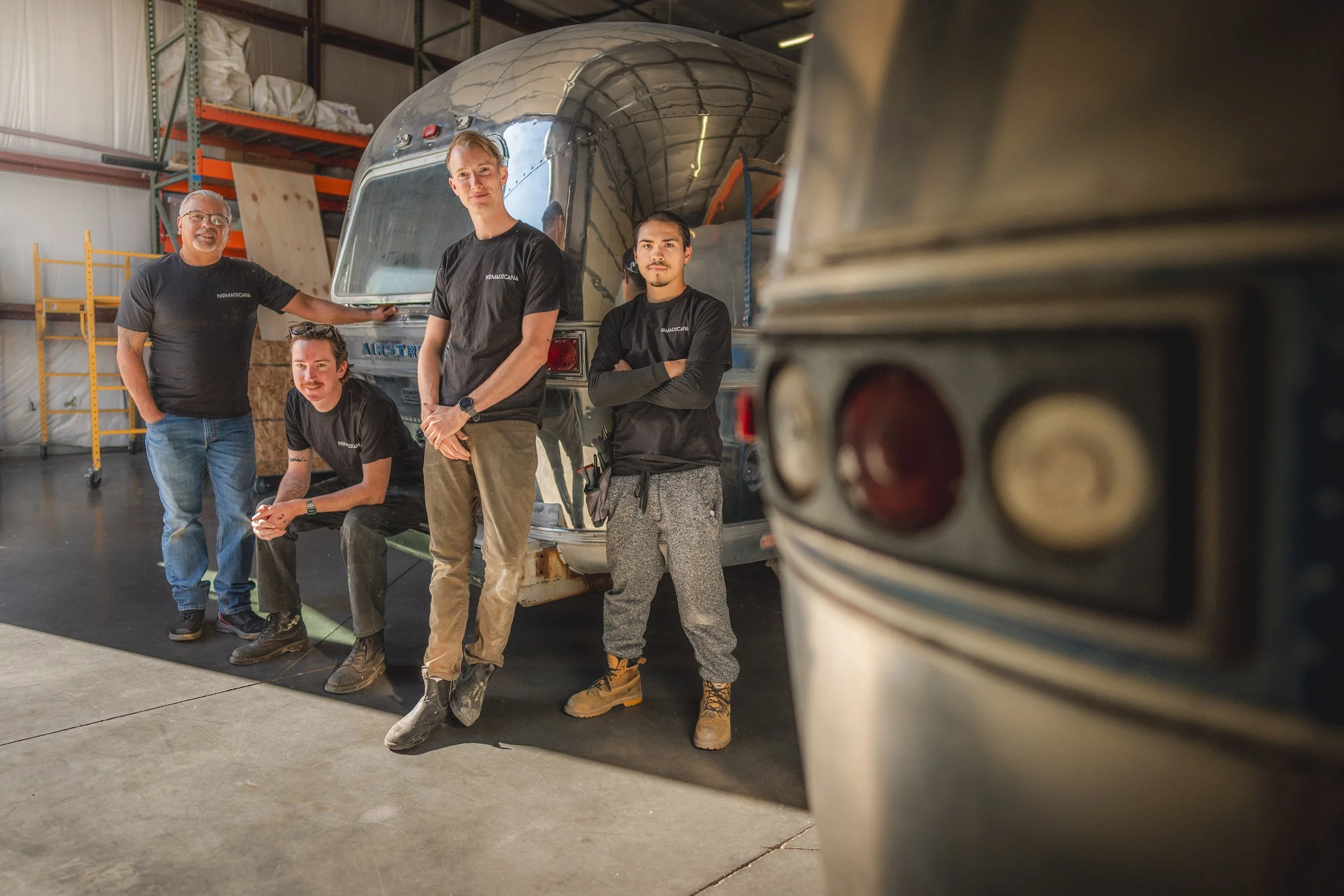 Five men standing and sitting near a shiny vintage trailer in an industrial warehouse, with another trailer partially visible in the foreground.
