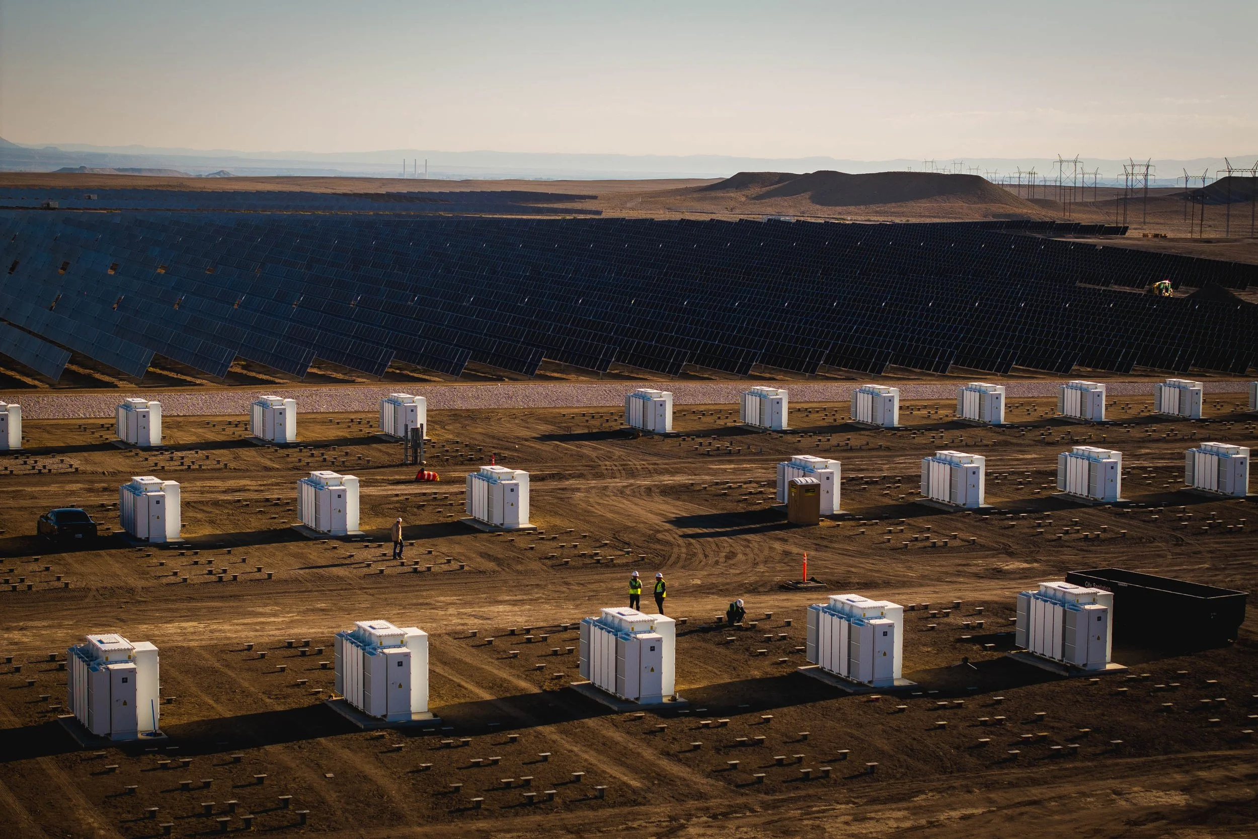 A large solar farm with numerous solar panels and small white structures on a dirt lot, with workers in safety vests.