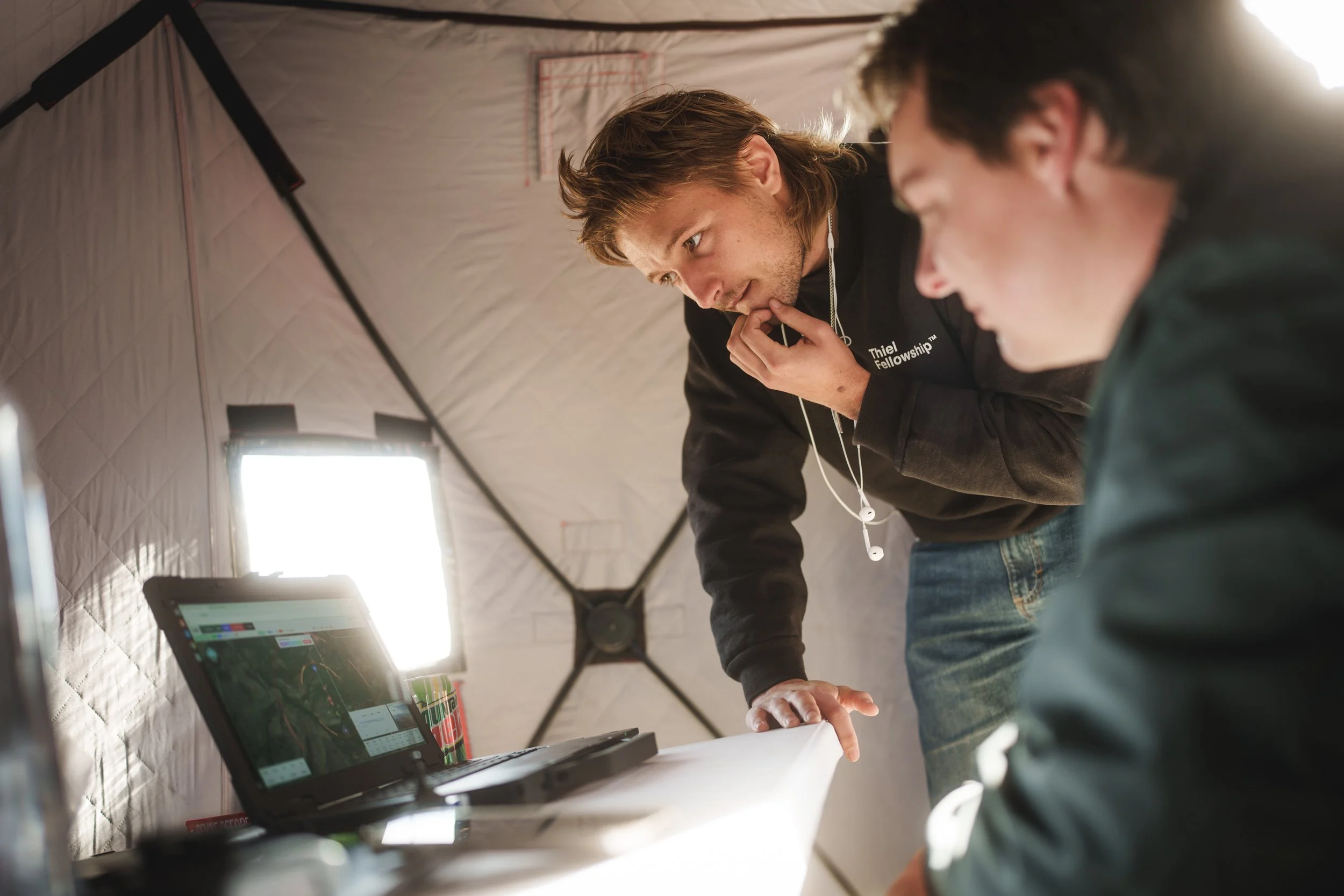 Two young men working on a laptop inside a tent, with one leaning over and looking intently at the screen and the other sitting nearby. Bright light source behind them, illuminating their workspace.