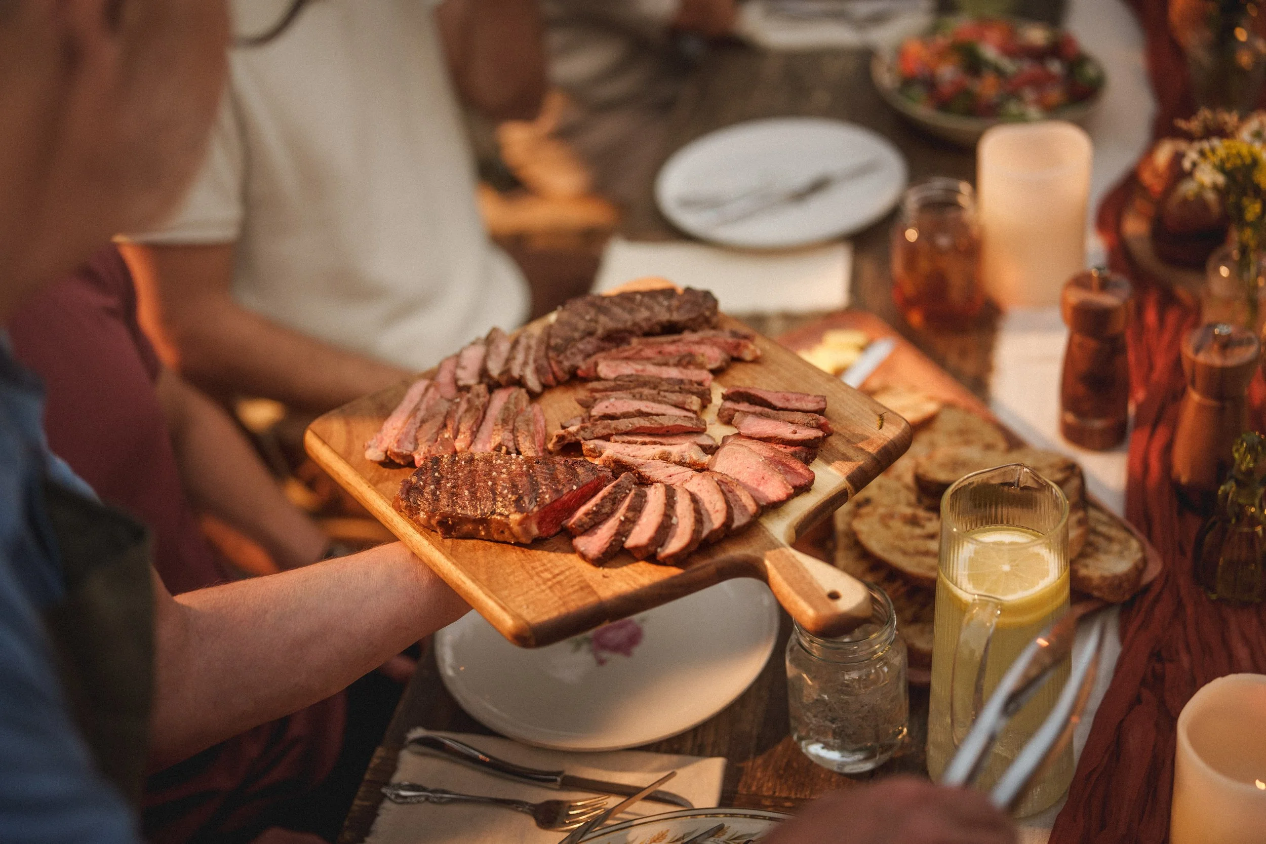 Person holding a wooden board with various sliced grilled meats at a dinner party table with food, drinks, and candles.