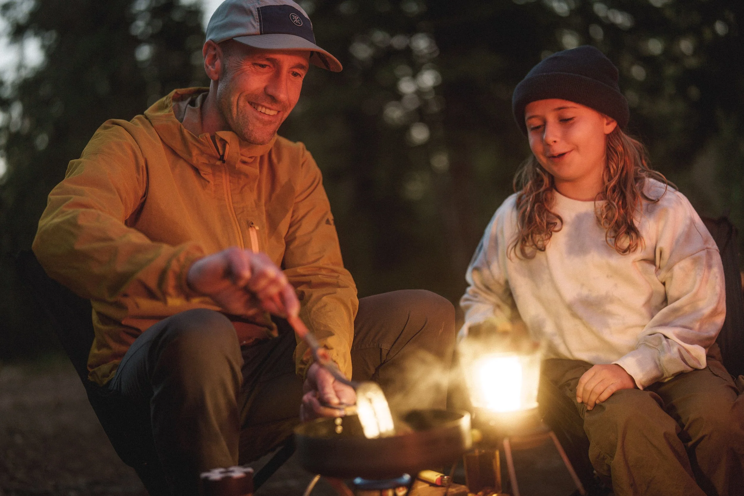 A man and girl sitting around a campfire at night, roasting marshmallows.
