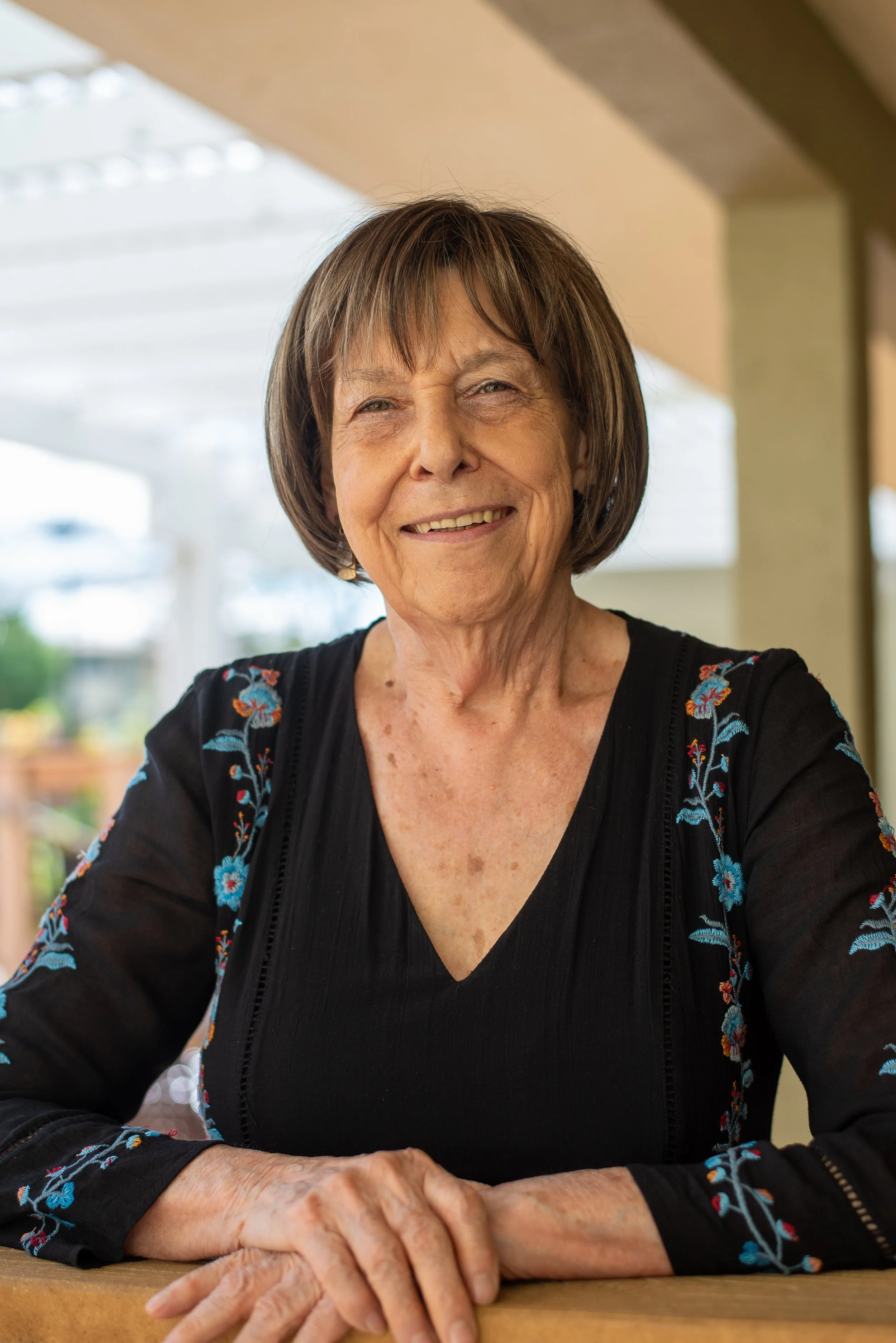 A smiling elderly woman with short brown hair sitting at a table indoors.