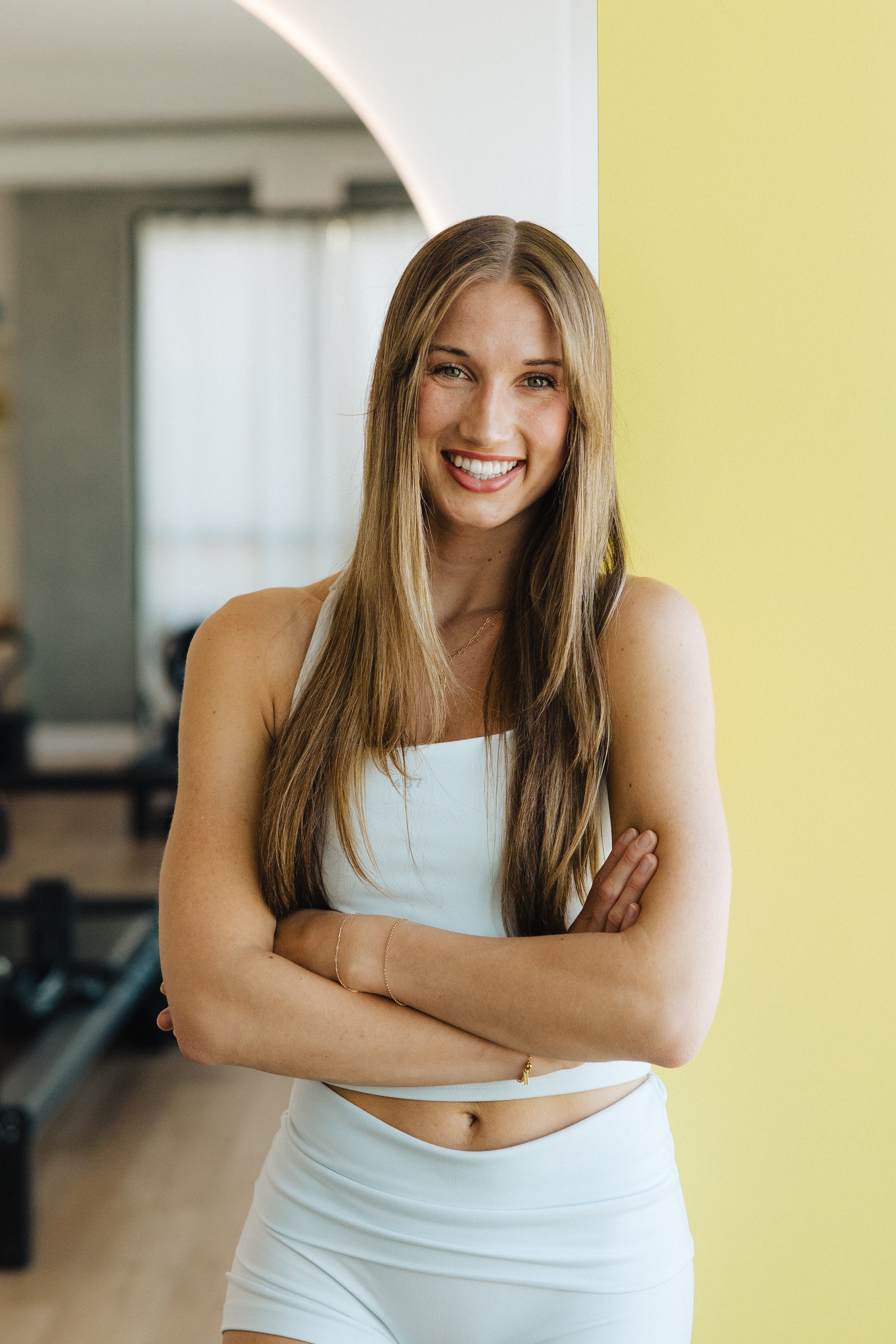 A smiling young woman with long blonde hair, wearing a black sleeveless top and light-colored pants, standing with arms crossed inside a modern indoor space with large windows and green and yellow walls.