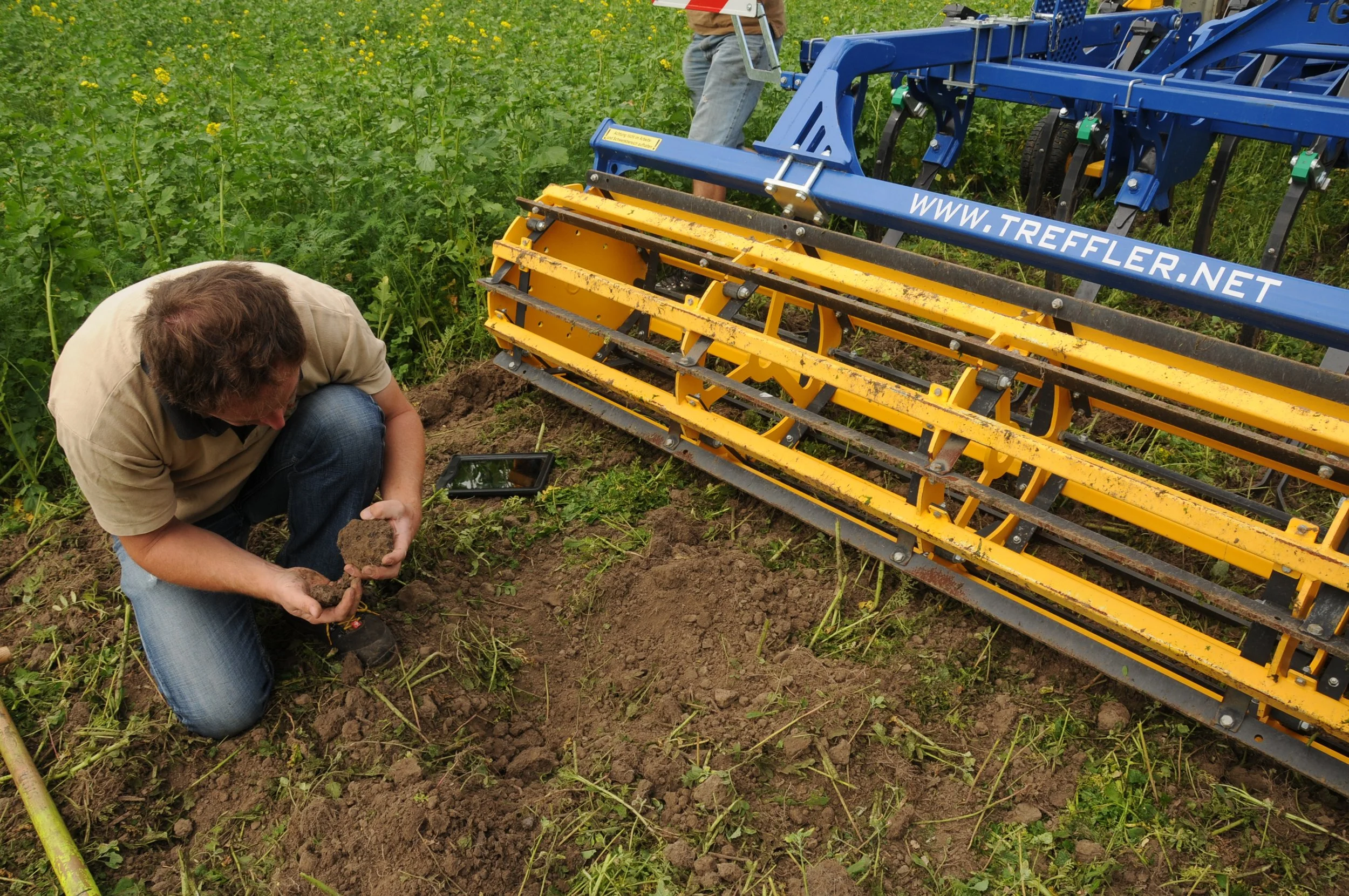 Un homme plie à genoux dans un champ de légumes, tenant une motte de terre tandis qu'une machine agricole jaune et bleue est positionnée à proximité pour le travail du sol.