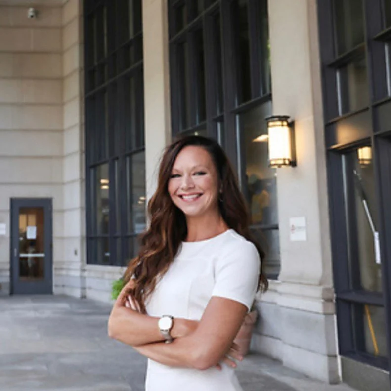 Woman in white dress smiling and standing with arms crossed outside a building with large windows.
