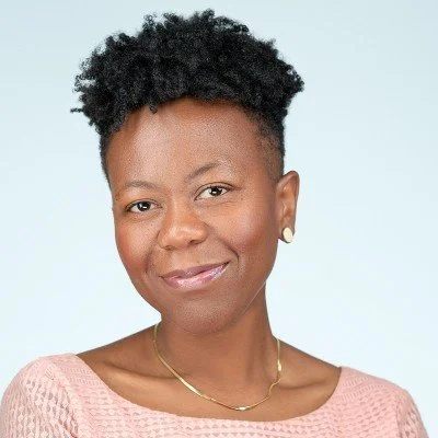 Portrait of a person with short curly hair, wearing a pink top and a gold necklace, against a light blue background.