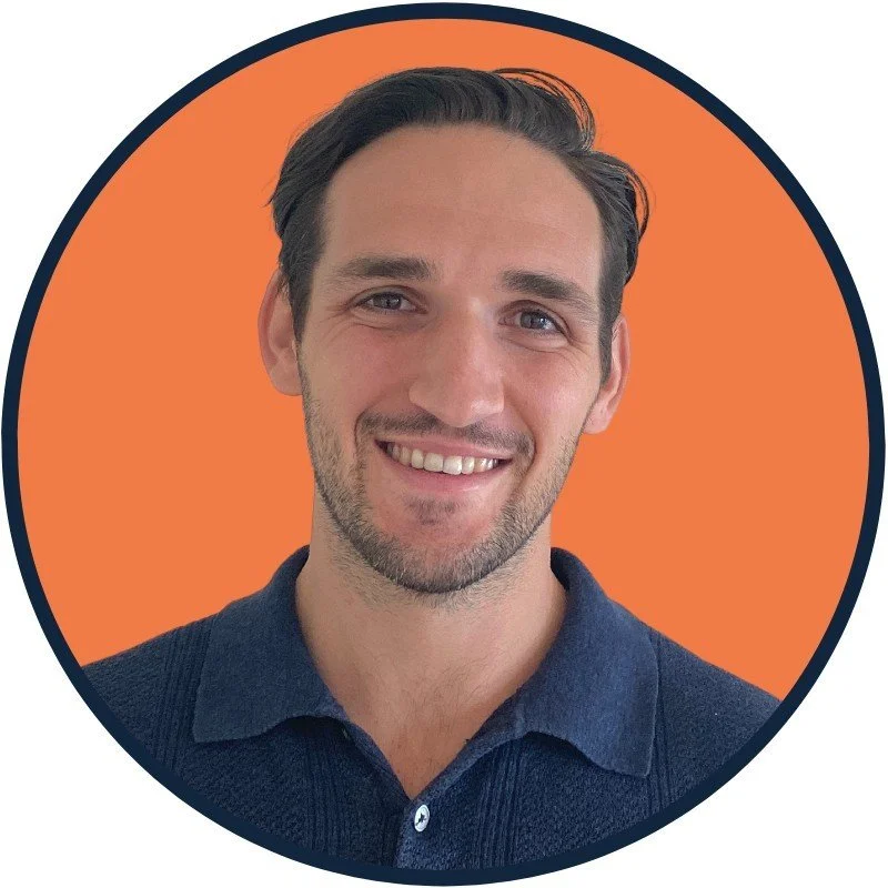 Smiling man with dark hair and beard, wearing blue collared shirt, against orange circular background.