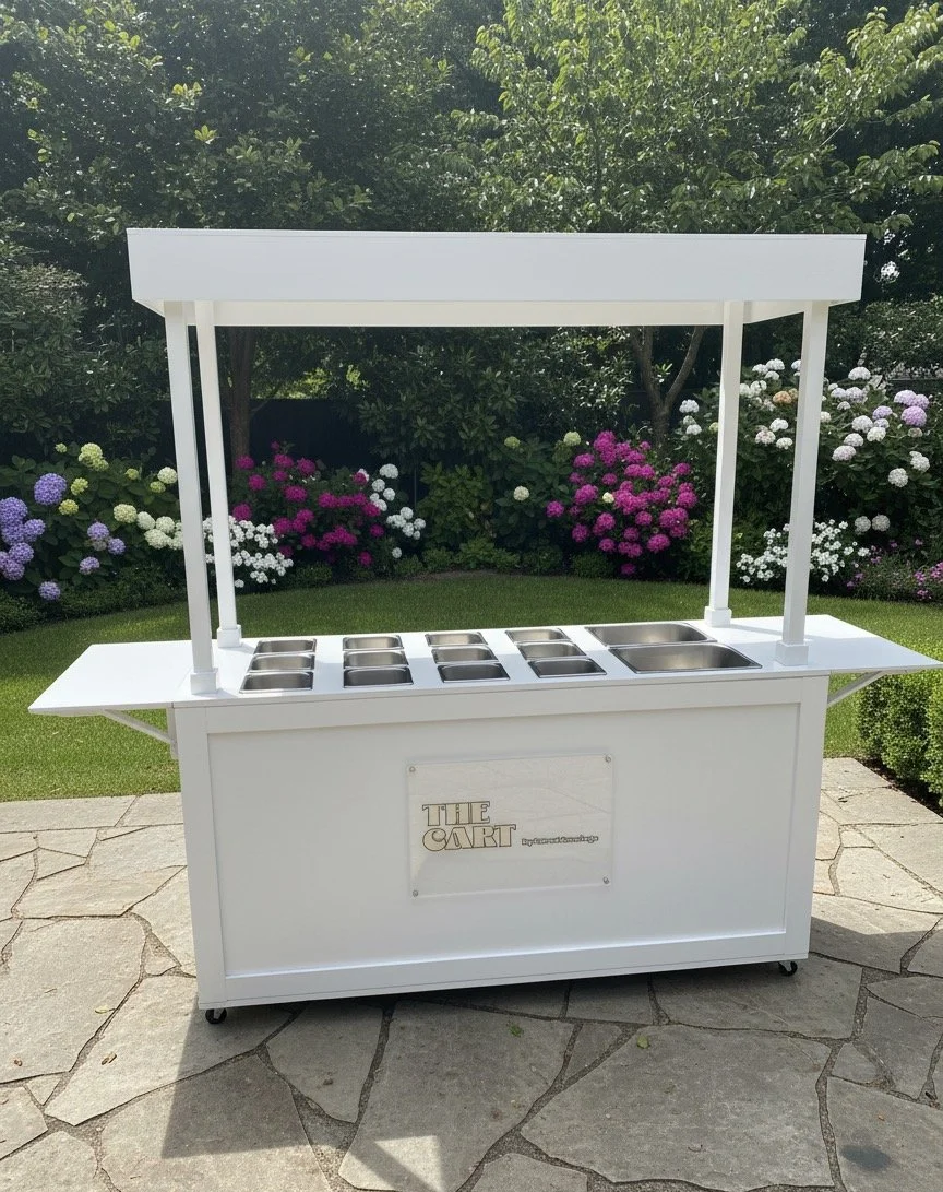White outdoor food cart with multiple stainless steel containers on top, situated on a stone patio with lush green bushes and colorful flowers in the background.