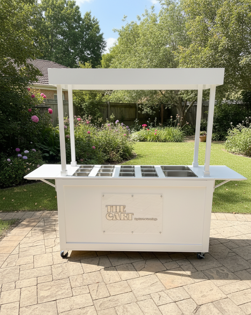 White mobile charcuterie food cart with a canopy, set outdoors on a paved surface, surrounded by green trees and flowering bushes.