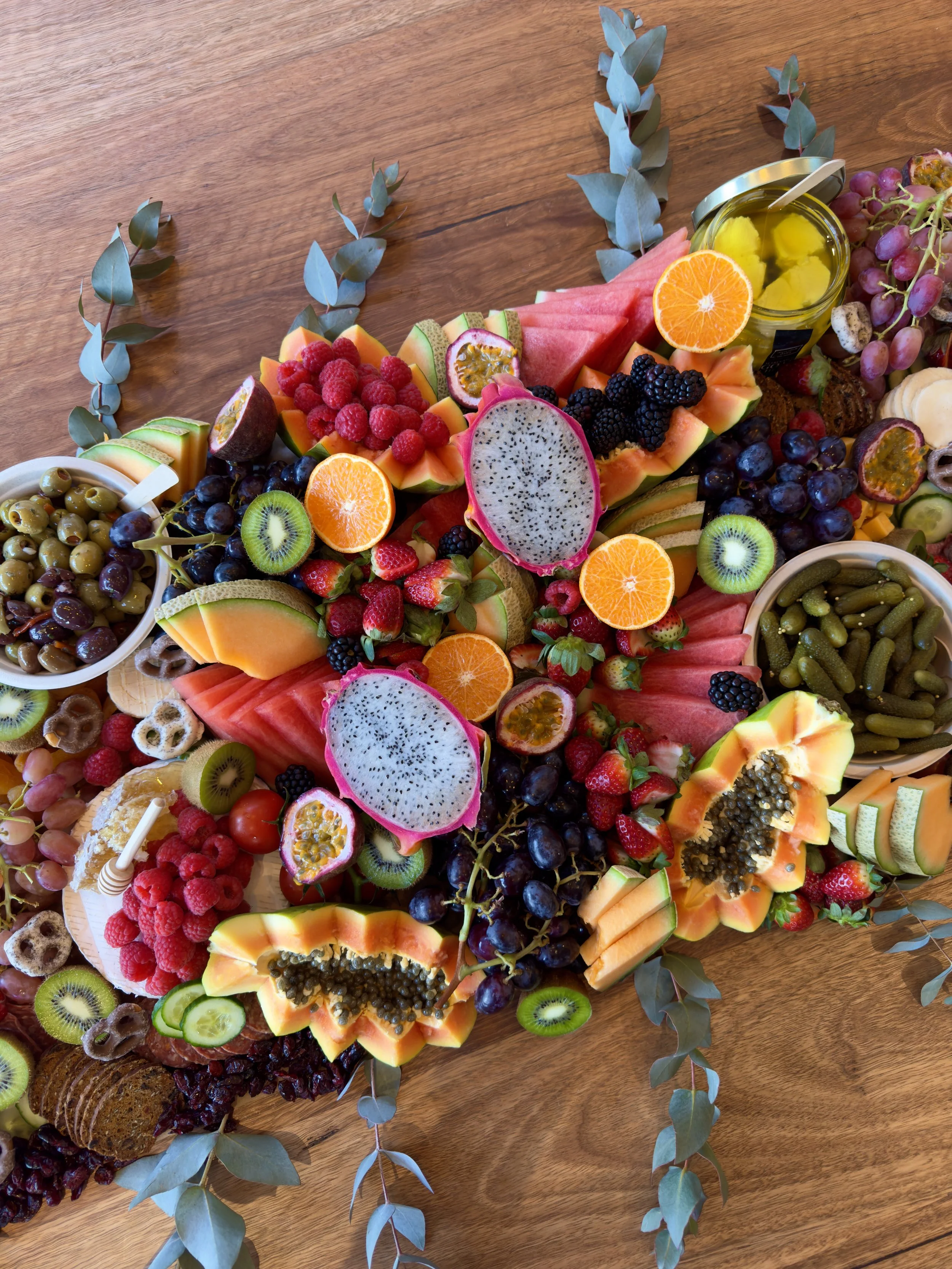 A colorful spread of various fresh fruits, including watermelon, cantaloupe, kiwi, dragon fruit, strawberries, raspberries, blackberries, grapes, and orange slices, arranged with some eucalyptus leaves on a wooden surface.