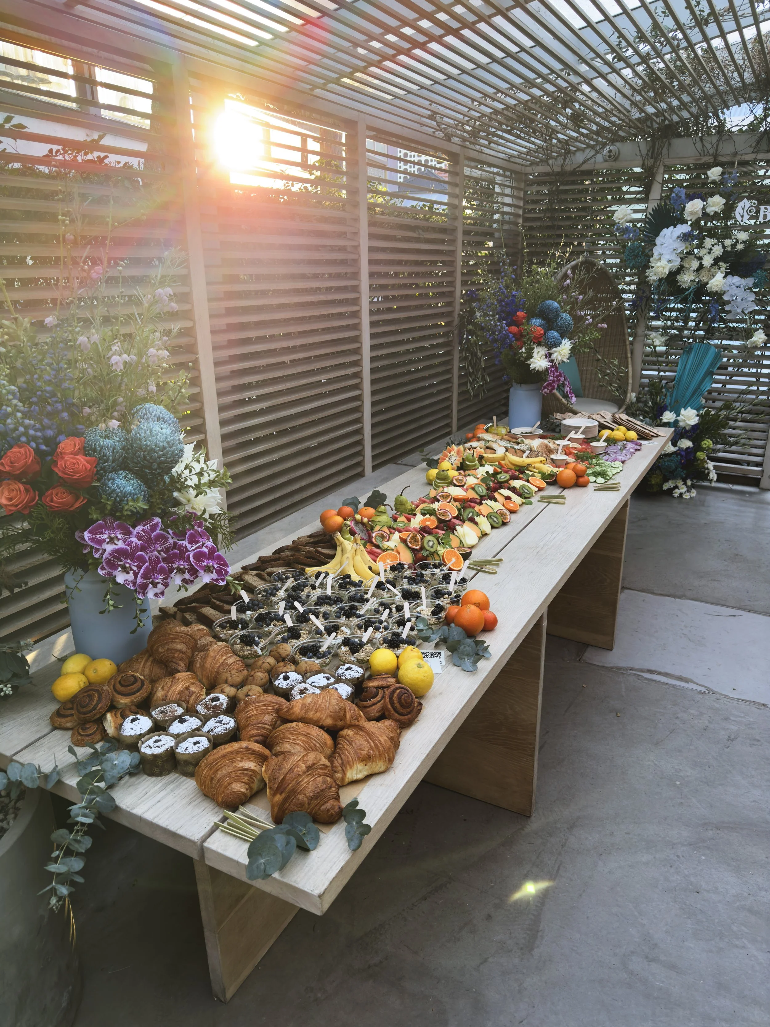 A long table with an assortment of desserts and fruit, decorated with colorful flowers in vases, set in a bright outdoor space with wooden slats and sunlight.