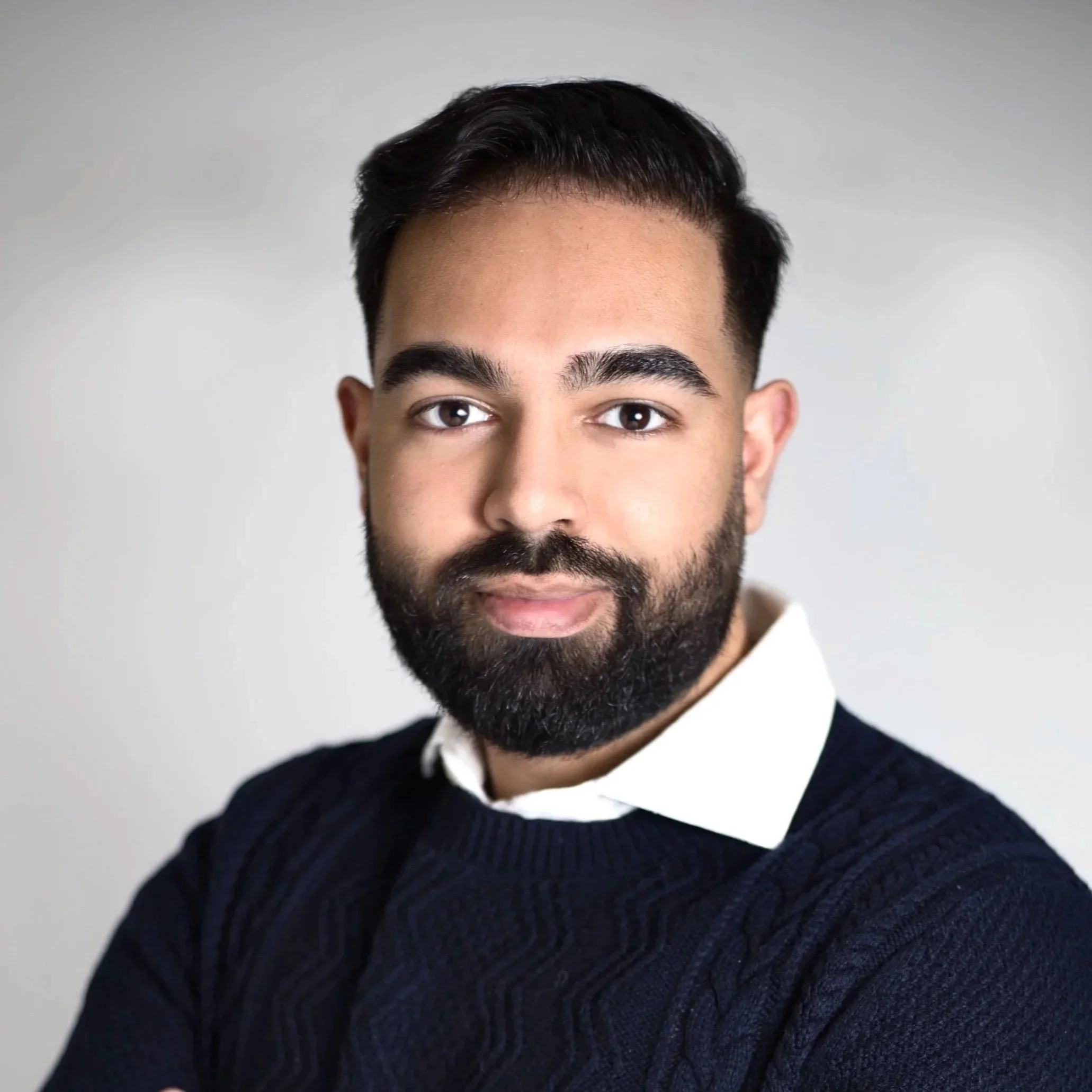 Male therapist with black hair and a black beard wearing a white collared shirt with a blue sweater smiling and looking straight