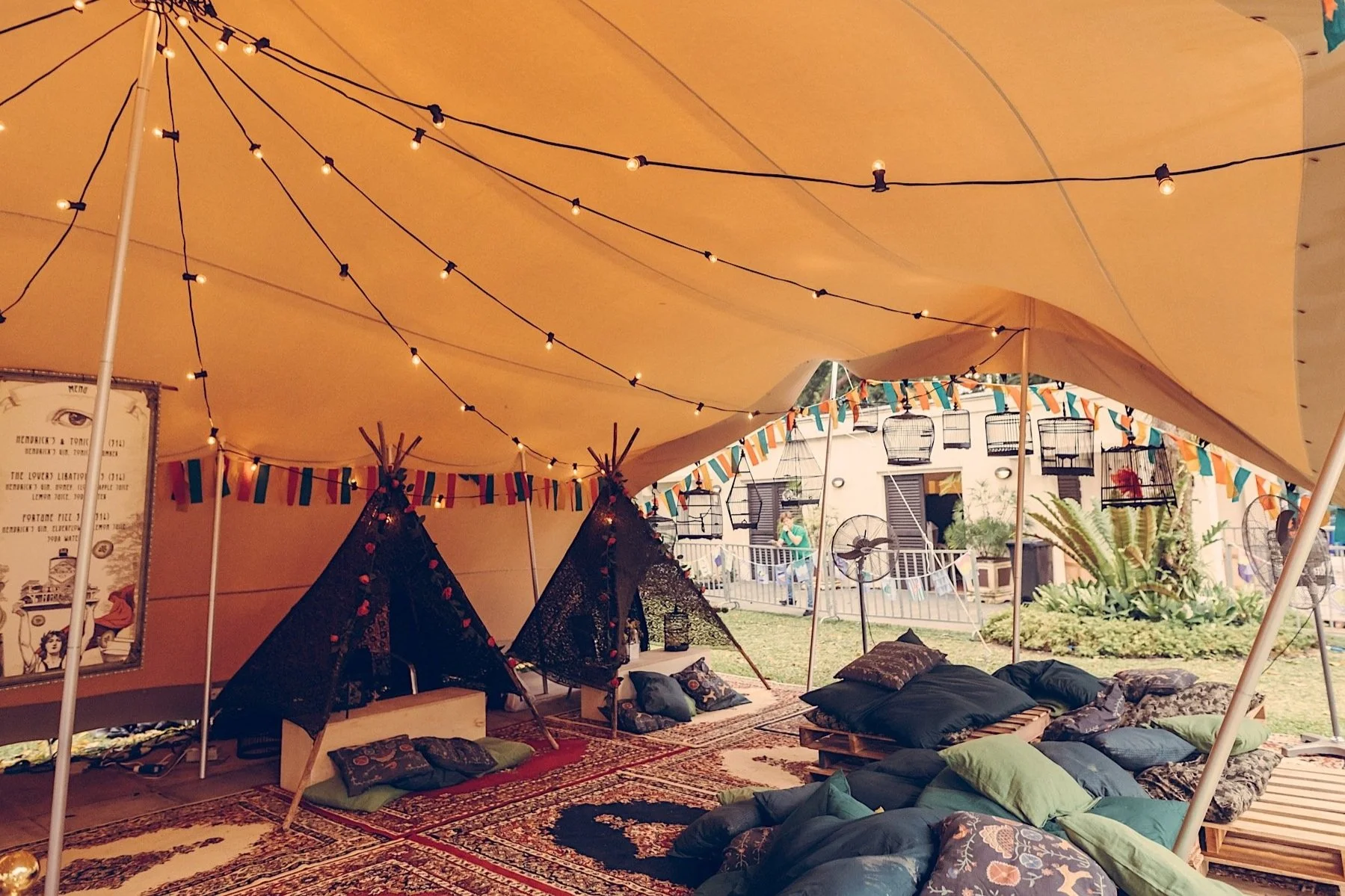 Interior of a large outdoor tent decorated with string lights, colorful bunting, and black lace teepees, with cushions and rugs on the ground for seating.