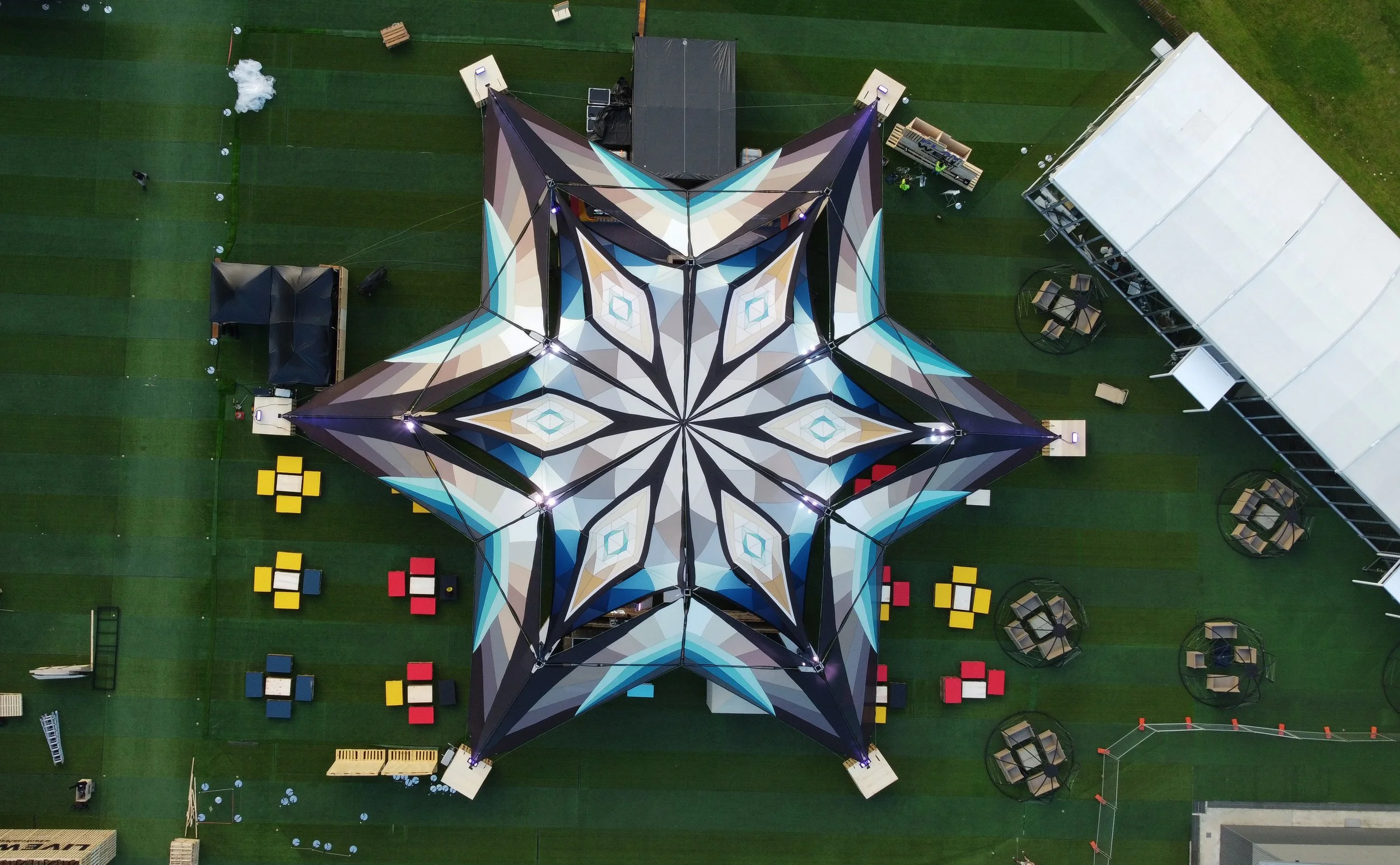 Aerial view of a large geometric canopy tent with a star-shaped pattern in shades of blue, beige, brown, and black, set up on a green artificial turf field with scattered seating and structures around it.