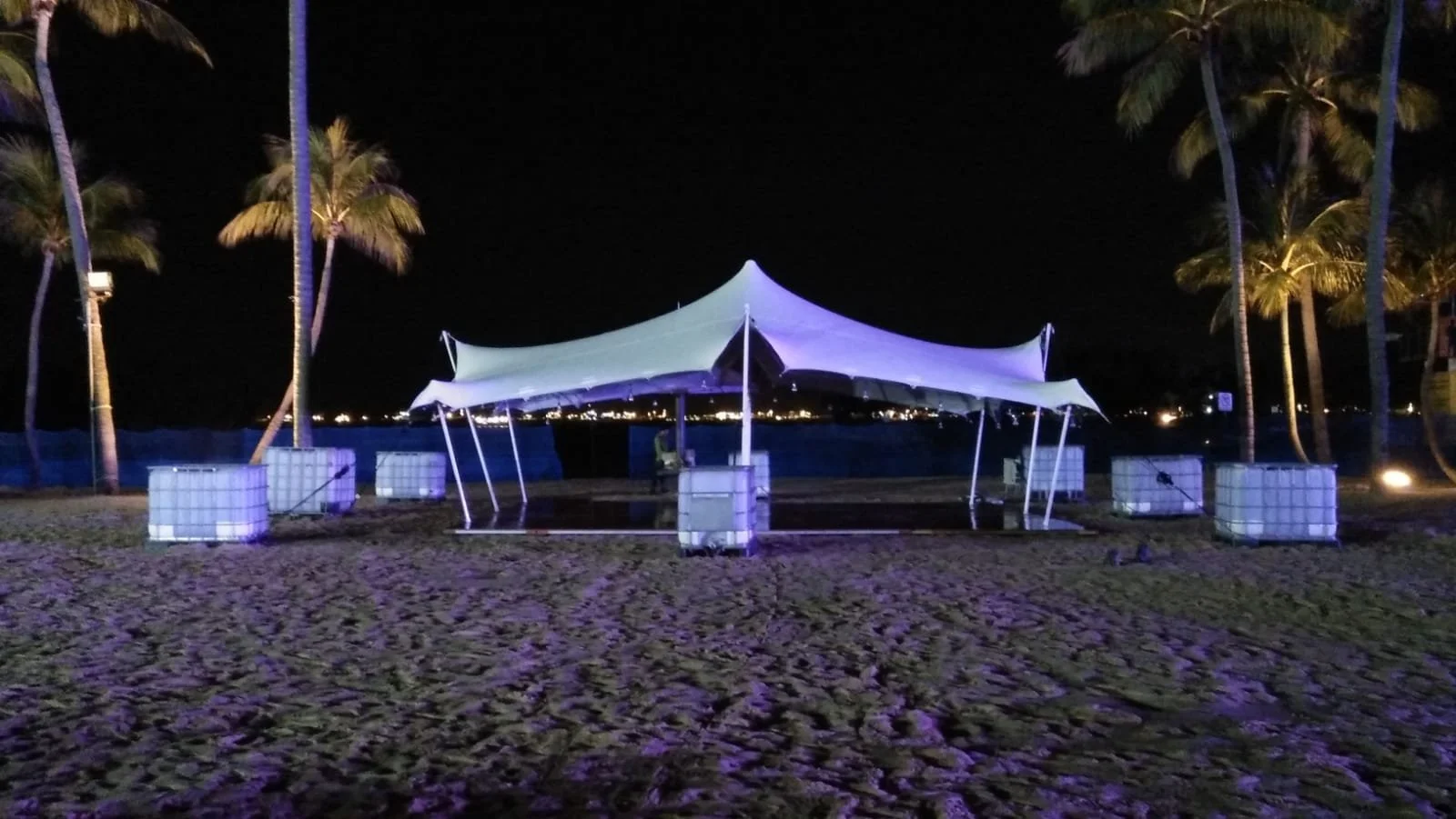 A nighttime scene on a beach featuring a large white canopy tent illuminated in purple lighting, palm trees, and floating cubes. In the background, city lights can be seen across the water.