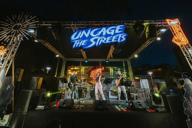 Live band performing on stage at night with a sign saying 'Uncage the Streets' and neon signs, including one that says 'Open.'