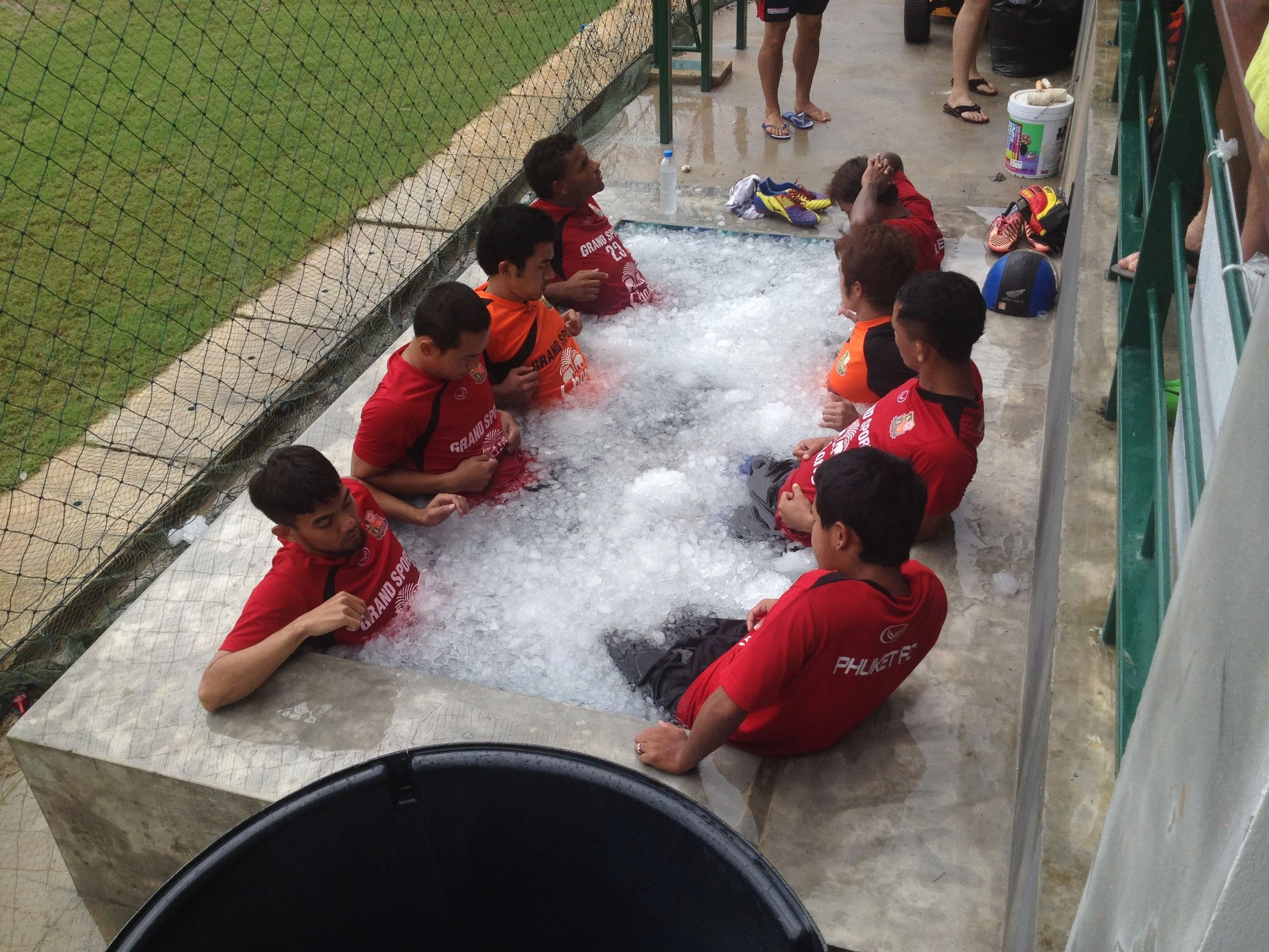 A football squad in red training tops sitting together in a pitchside pool of crushed ice after training