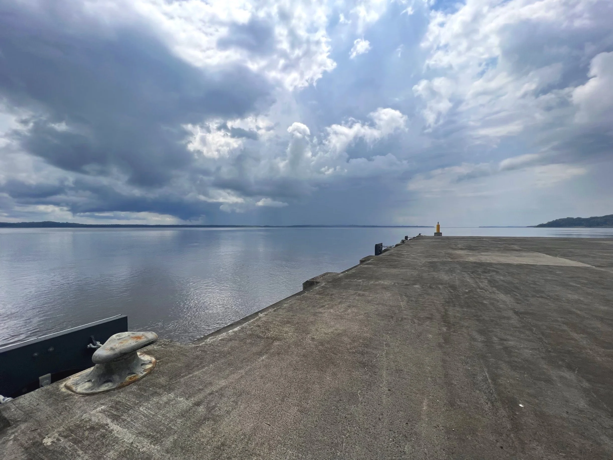 Concrete pier extending into a large body of calm water under a cloudy sky.