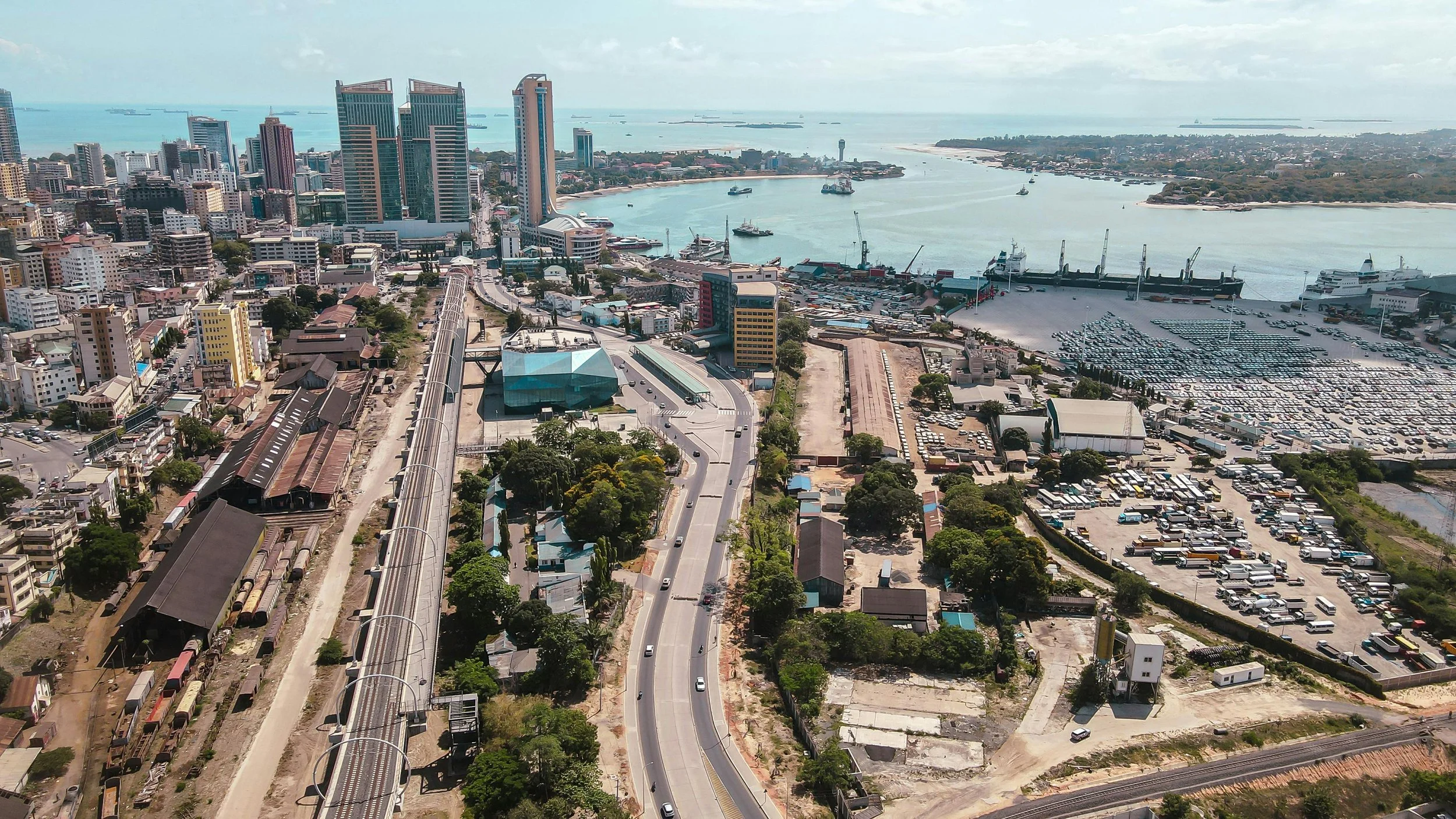 Aerial view of a cityscape with tall buildings, a port with docked ships, and parking lots filled with vehicles. A road and railway line run through the area, with greenery and industrial structures visible.