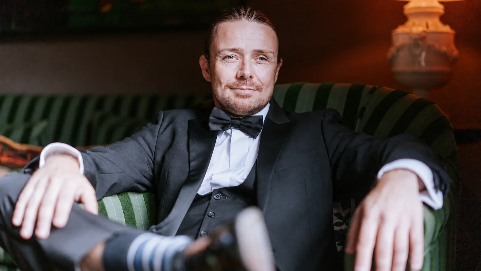 Just Saul, in a tuxedo, seated on a deep green sofa