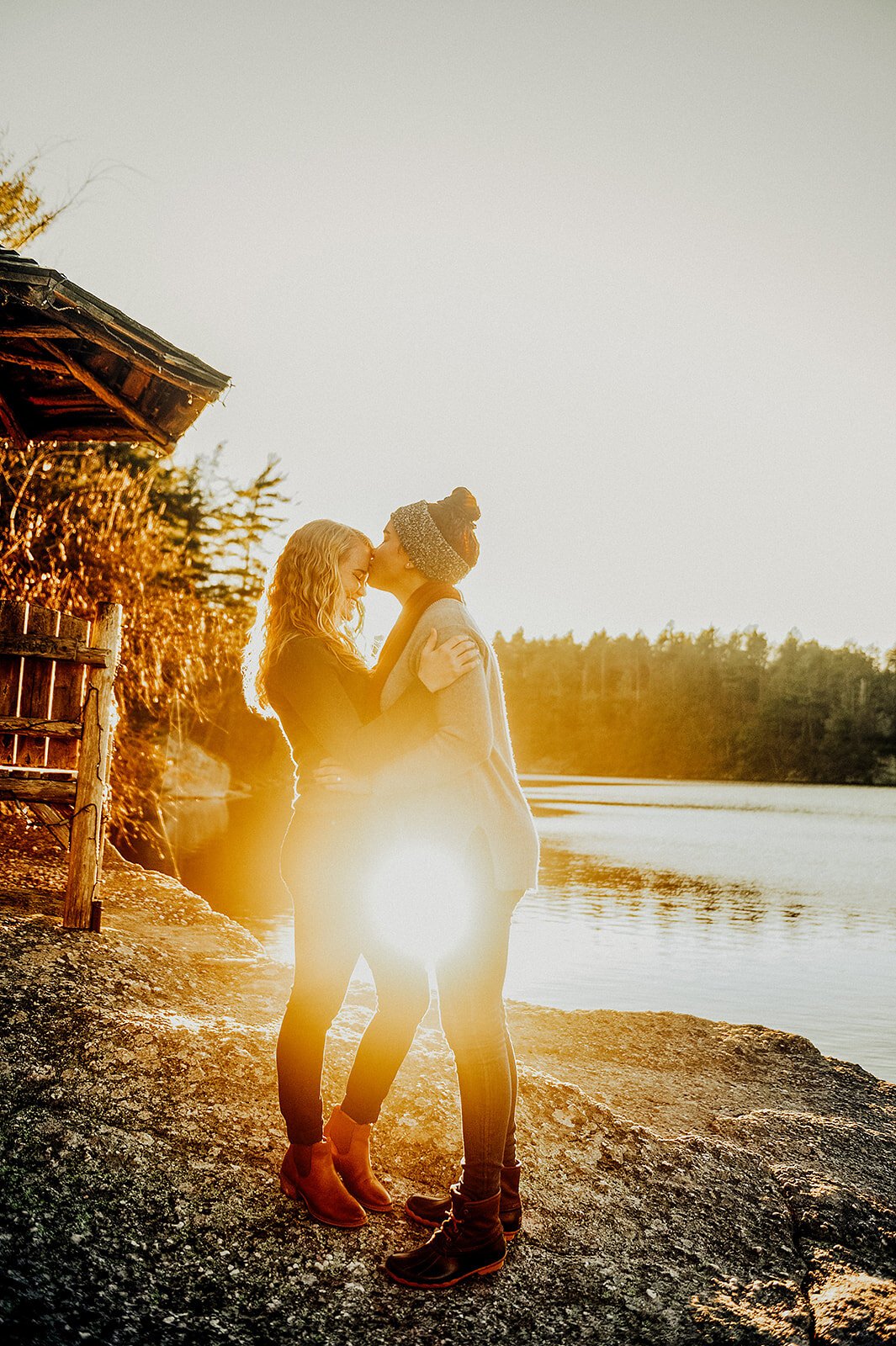 Two women sharing a kiss on a rocky shoreline at sunset, with a body of water and trees in the background.