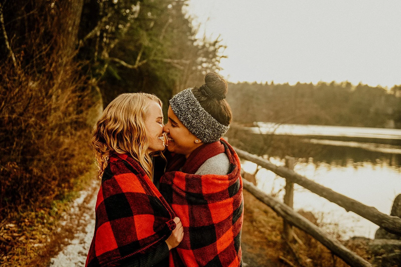 Two women outside by a lake, wrapped in red and black plaid blankets, smiling and touching foreheads during sunset.