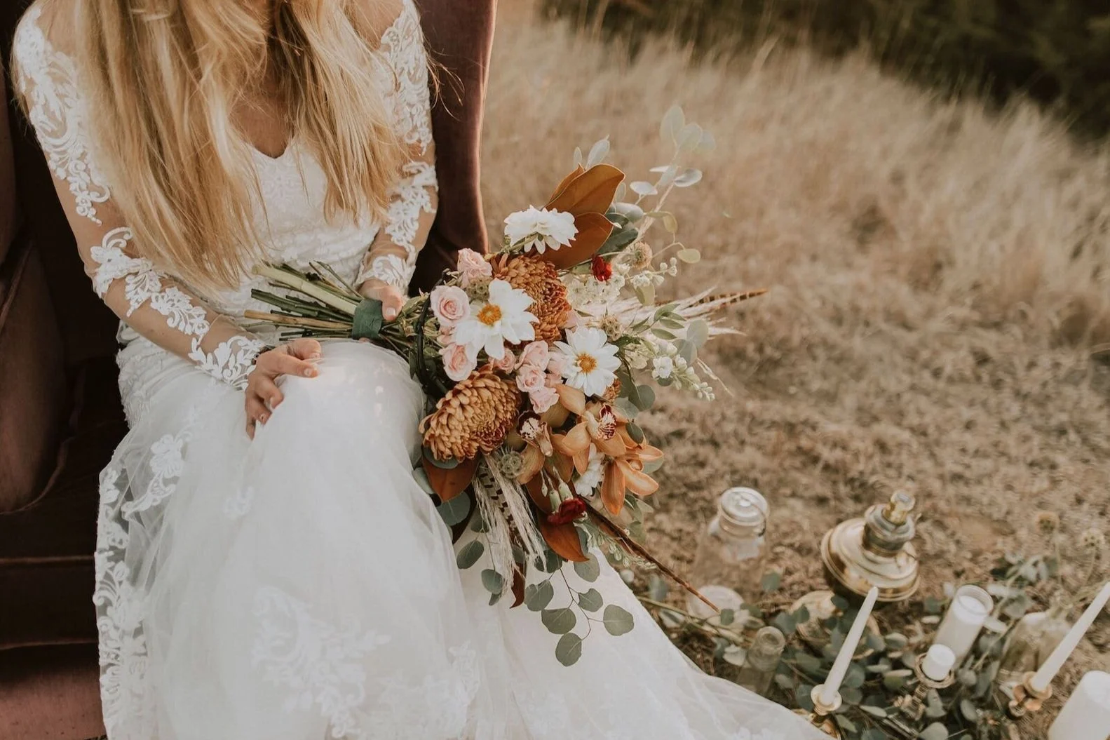 Bride in a lace wedding dress holding a bouquet of mixed flowers, sitting on a vintage sofa outdoors near candles and lanterns.