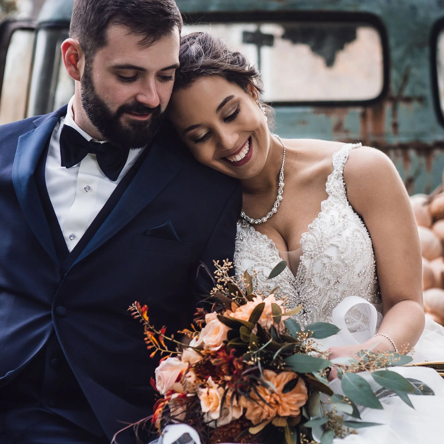 A newlywed couple sits close together, smiling, with the bride holding a bouquet of peach, orange, and green flowers, while leaning their heads against a rustic, vintage vehicle in the background.
