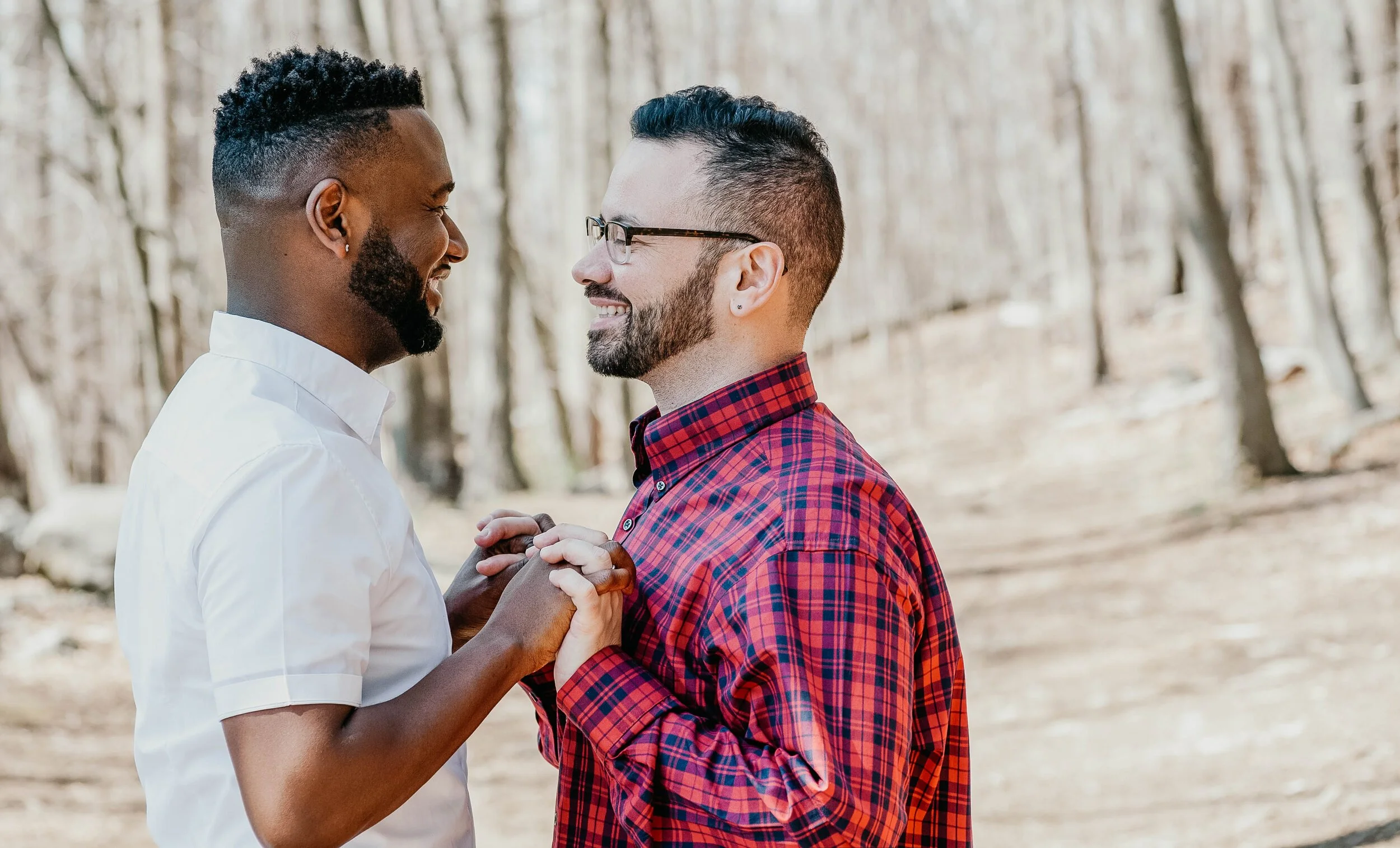 Two men smiling and holding hands in a wooded outdoor setting.