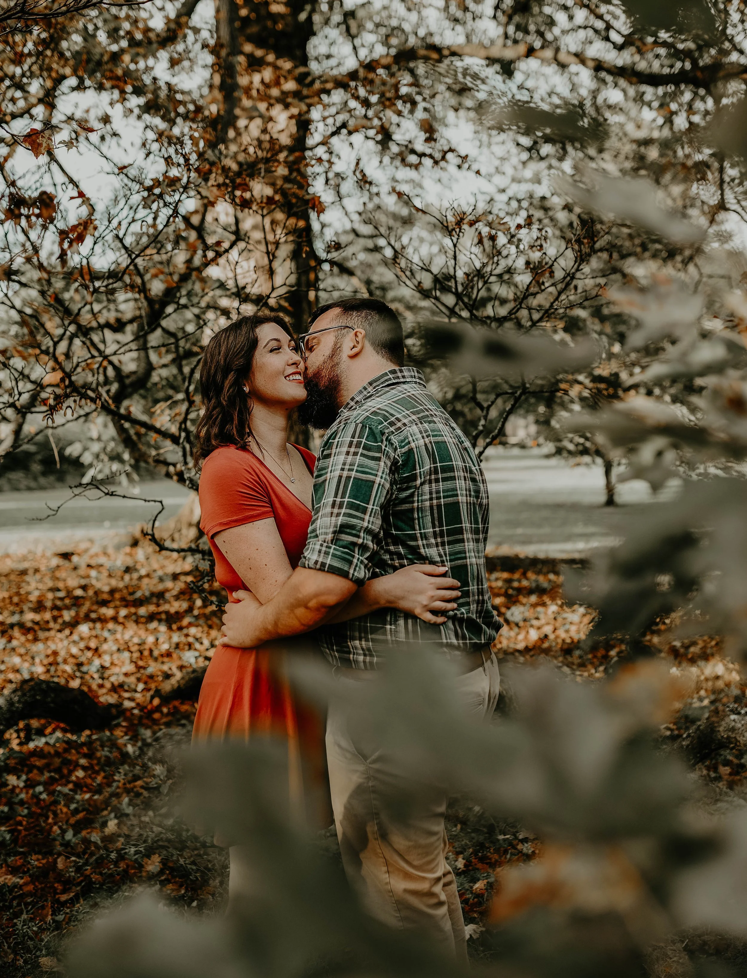 A couple standing close, about to kiss, outdoors among trees with autumn leaves, the woman in a red dress and the man in a plaid shirt.