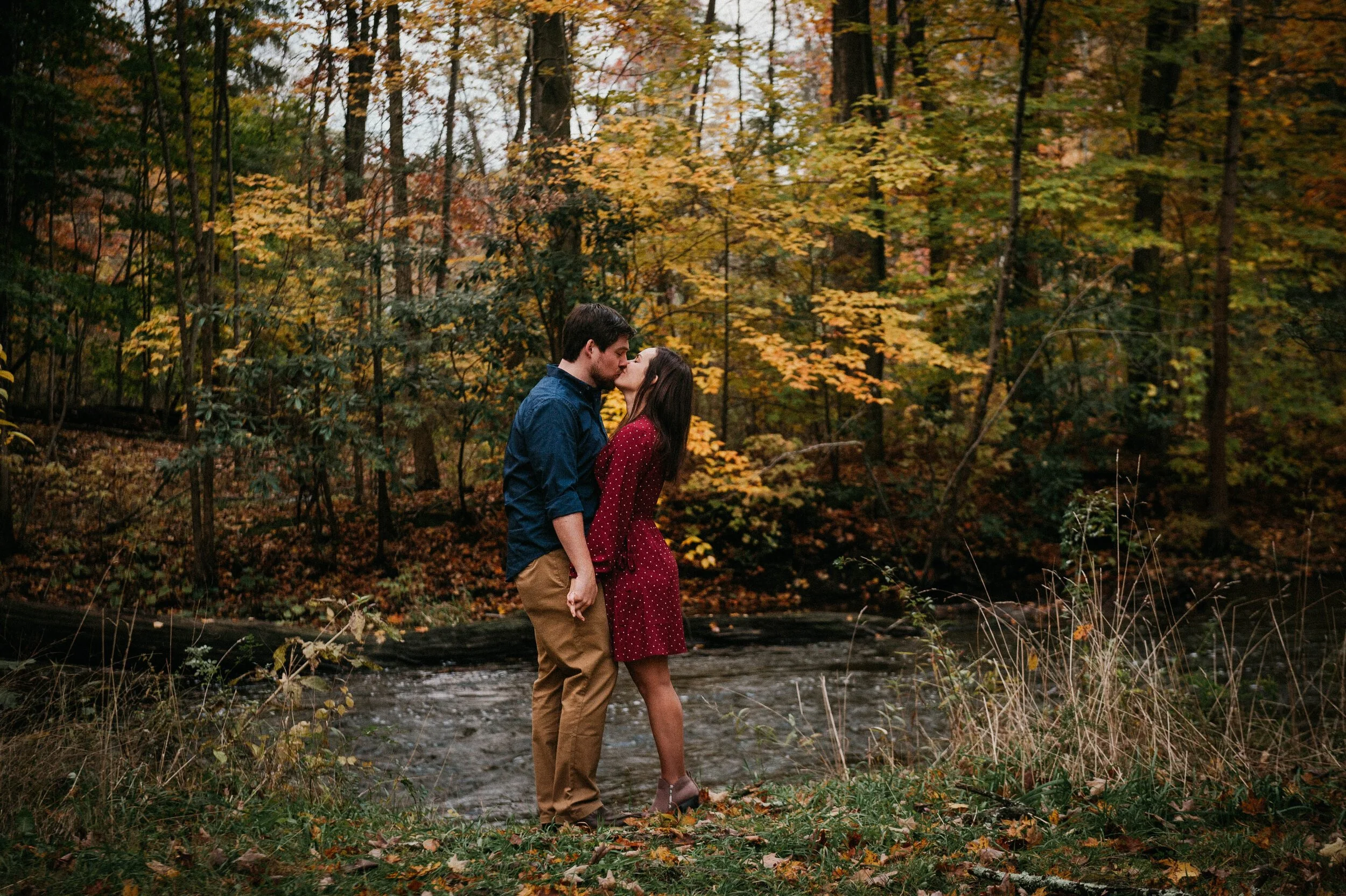 A couple kissing on a trail along a creek in a forest with autumn foliage.