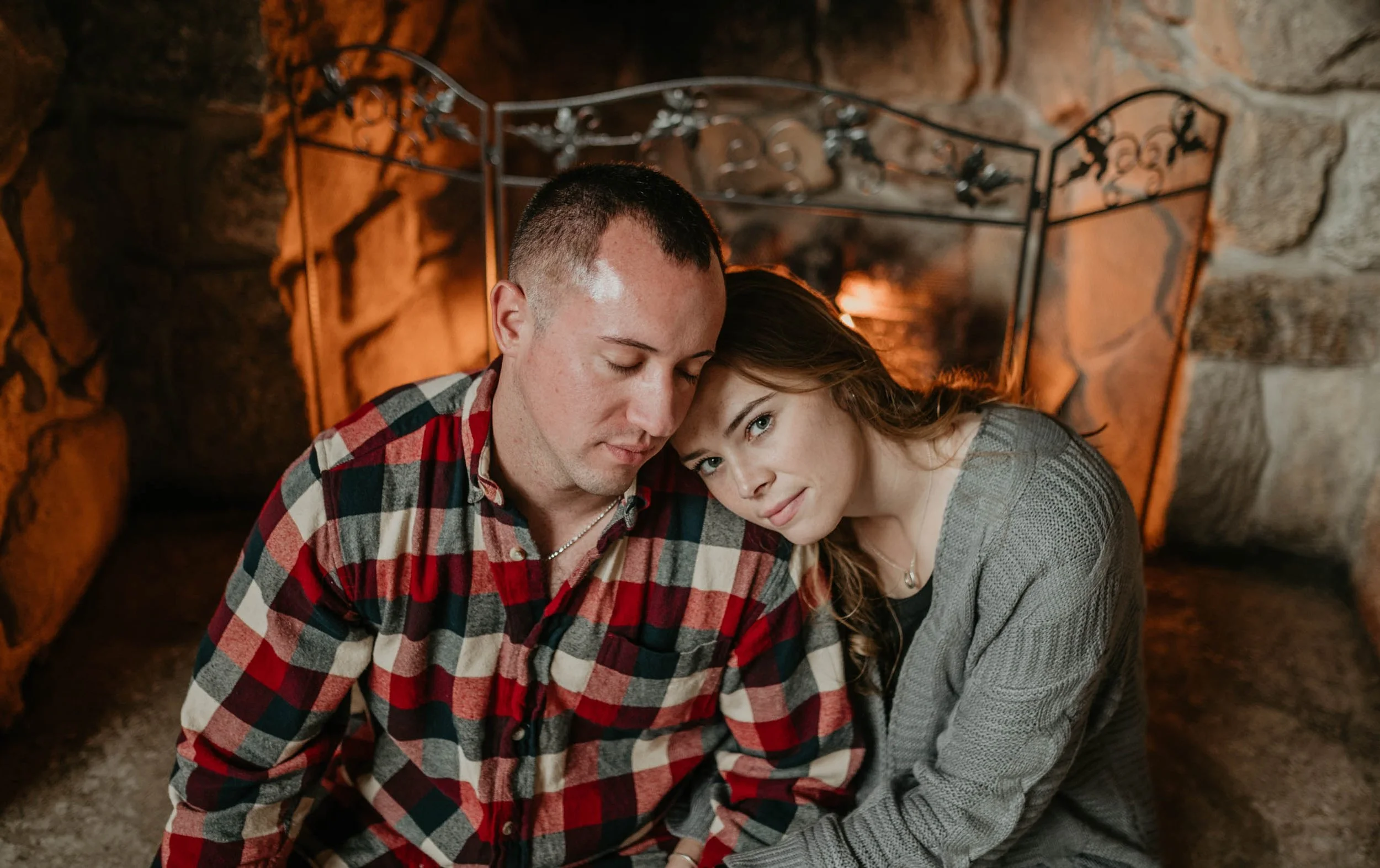 A couple sitting close together in front of a fireplace, with their foreheads touching and eyes closed, creating an intimate moment.
