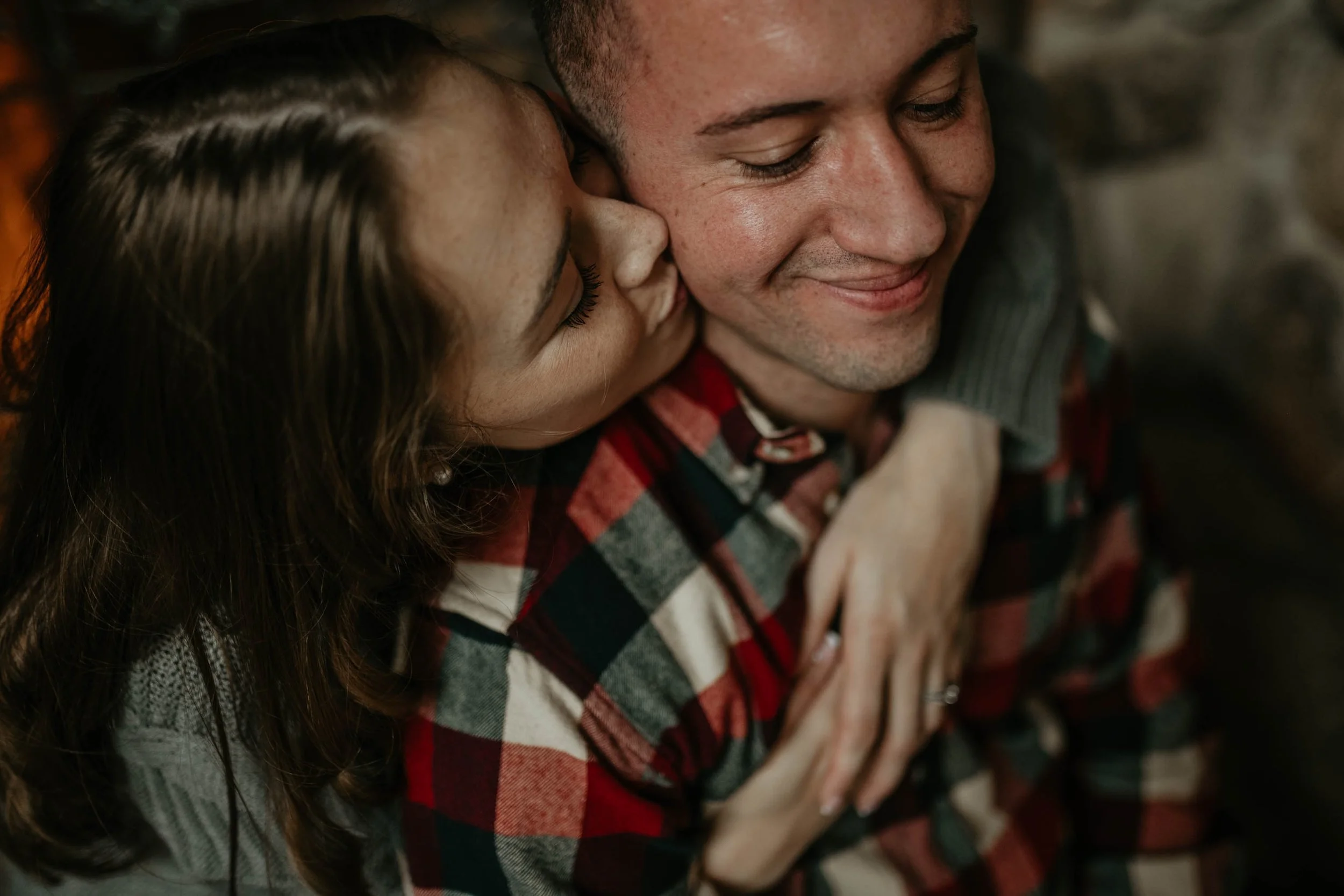 A woman with long brown hair kisses a man on the cheek. The man is smiling with his eyes closed, and the woman has her hand on the man's shoulder. The man is wearing a red, black, and white plaid shirt. The background is dimly lit.