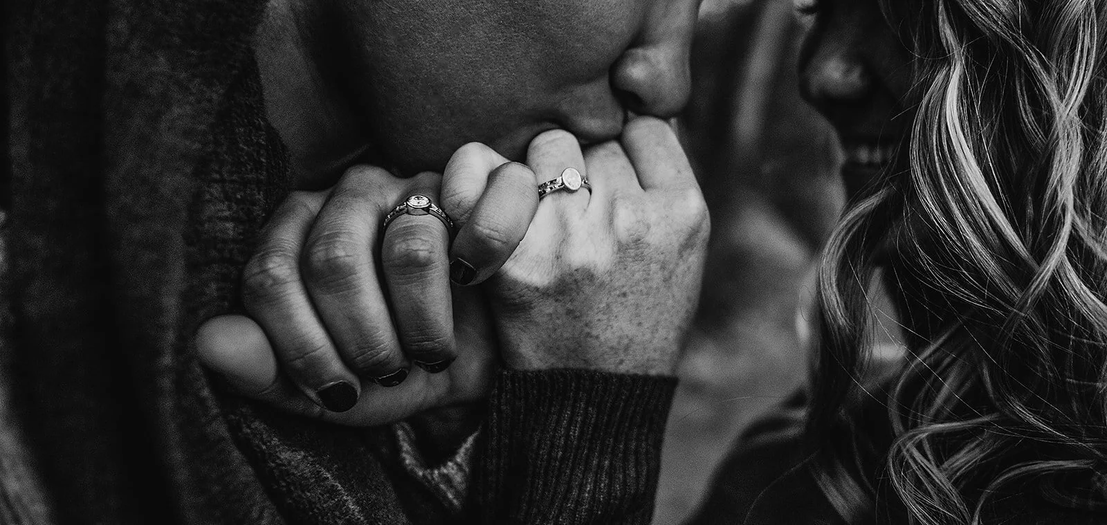 A close-up black and white photo of a person kissing another person's forehead, both with hands clasped together, and multiple rings on their fingers.