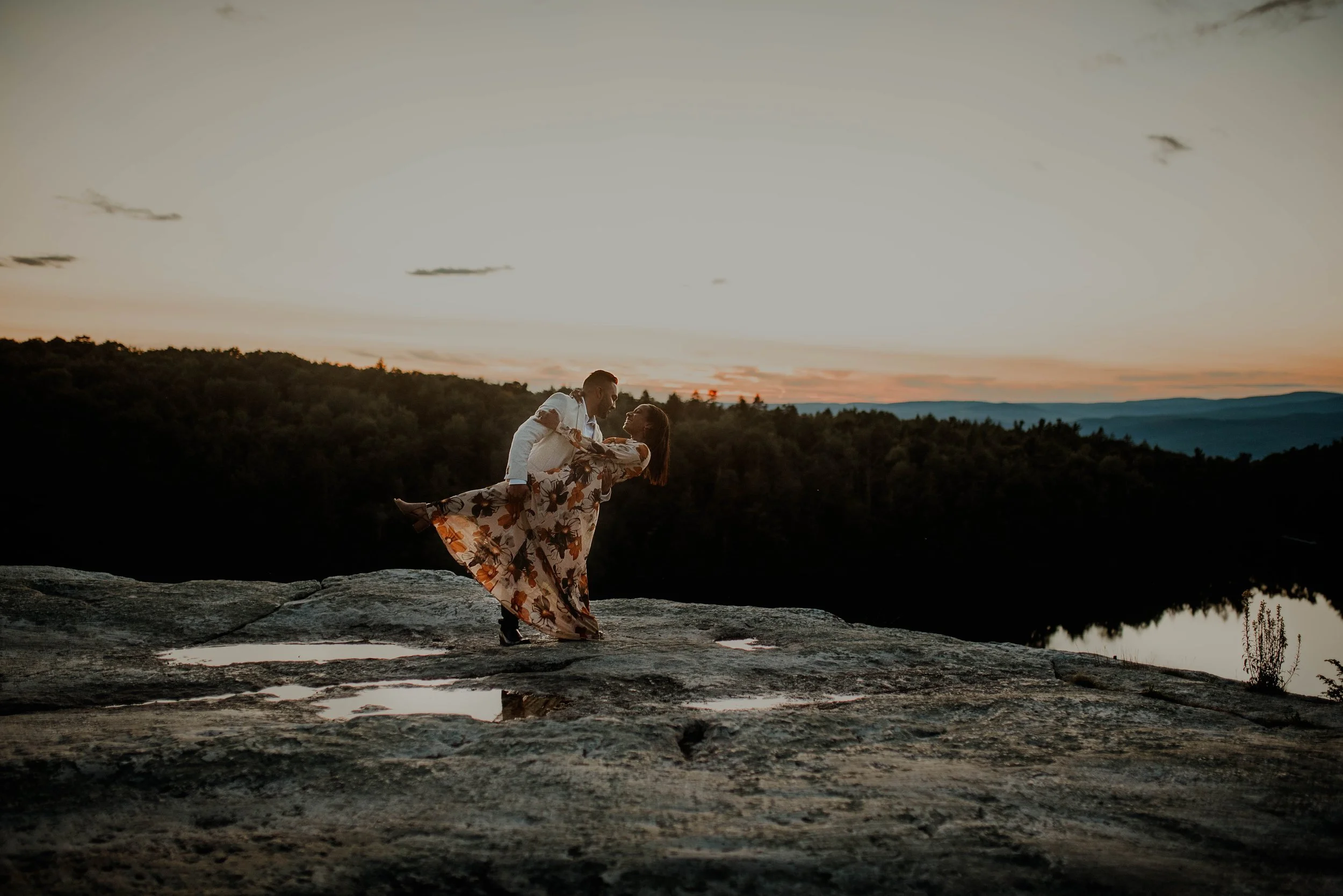 A couple dancing on a rocky ledge near a river at sunset, with a forested landscape and mountains in the background.