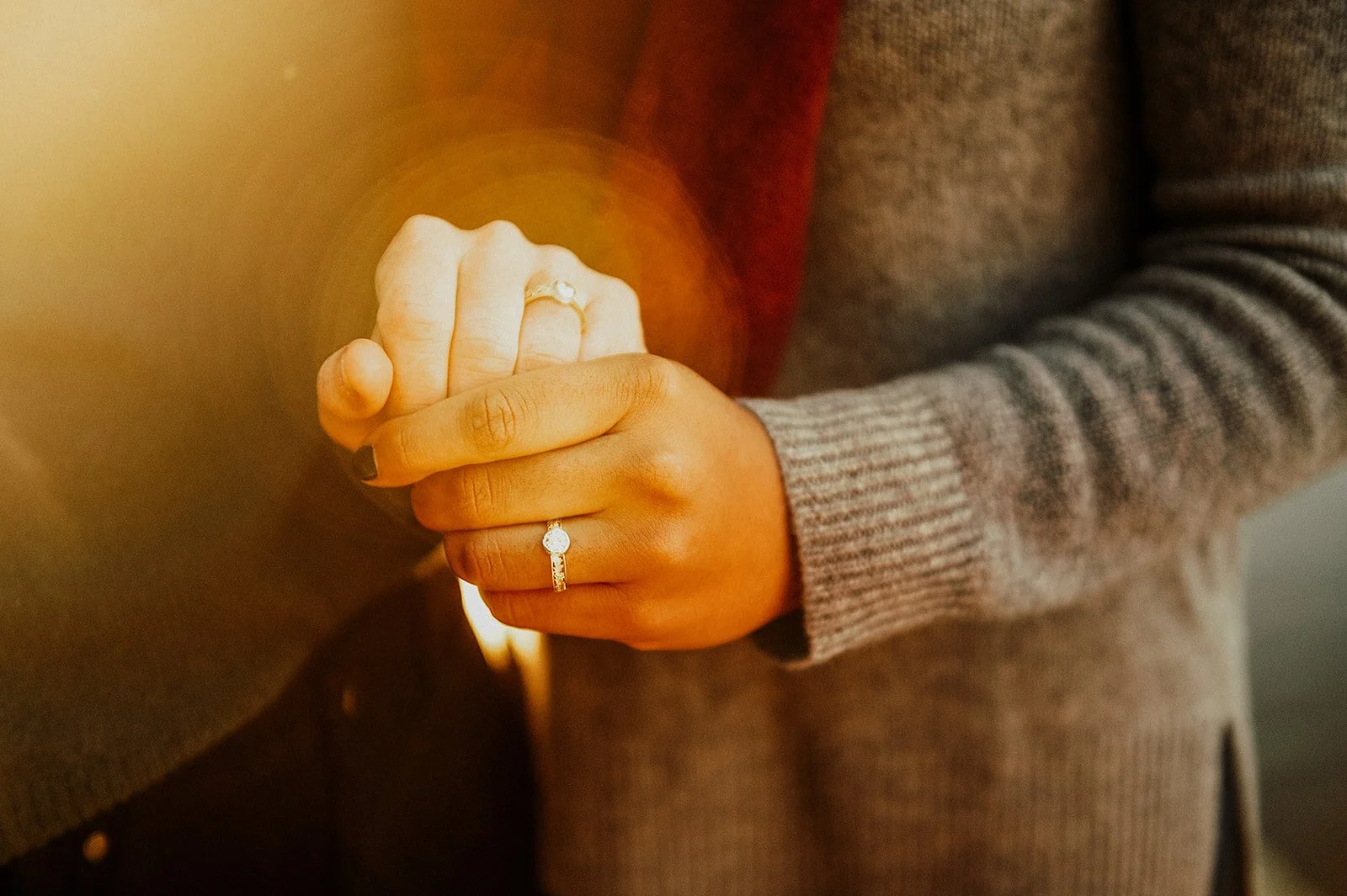 Close-up of two hands gently clasped, showcasing engagement rings, with warm lighting creating a soft glow.