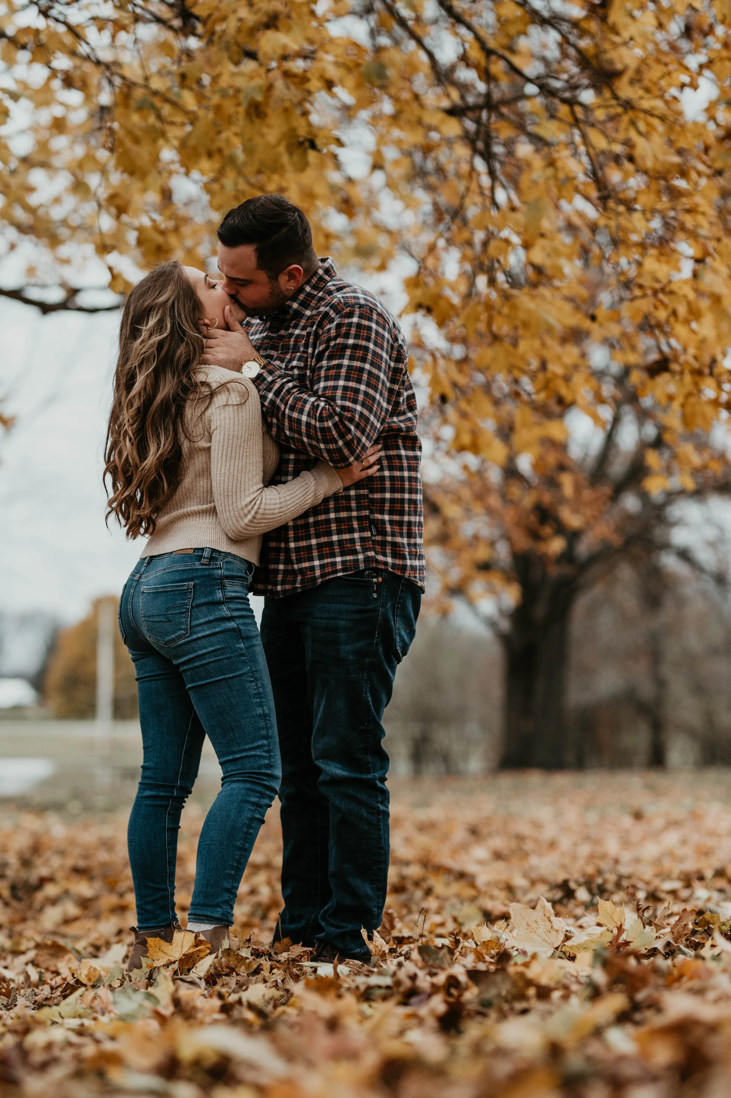 A couple kissing outdoors on a fall day, surrounded by orange and yellow leaves, with a large tree in the background.