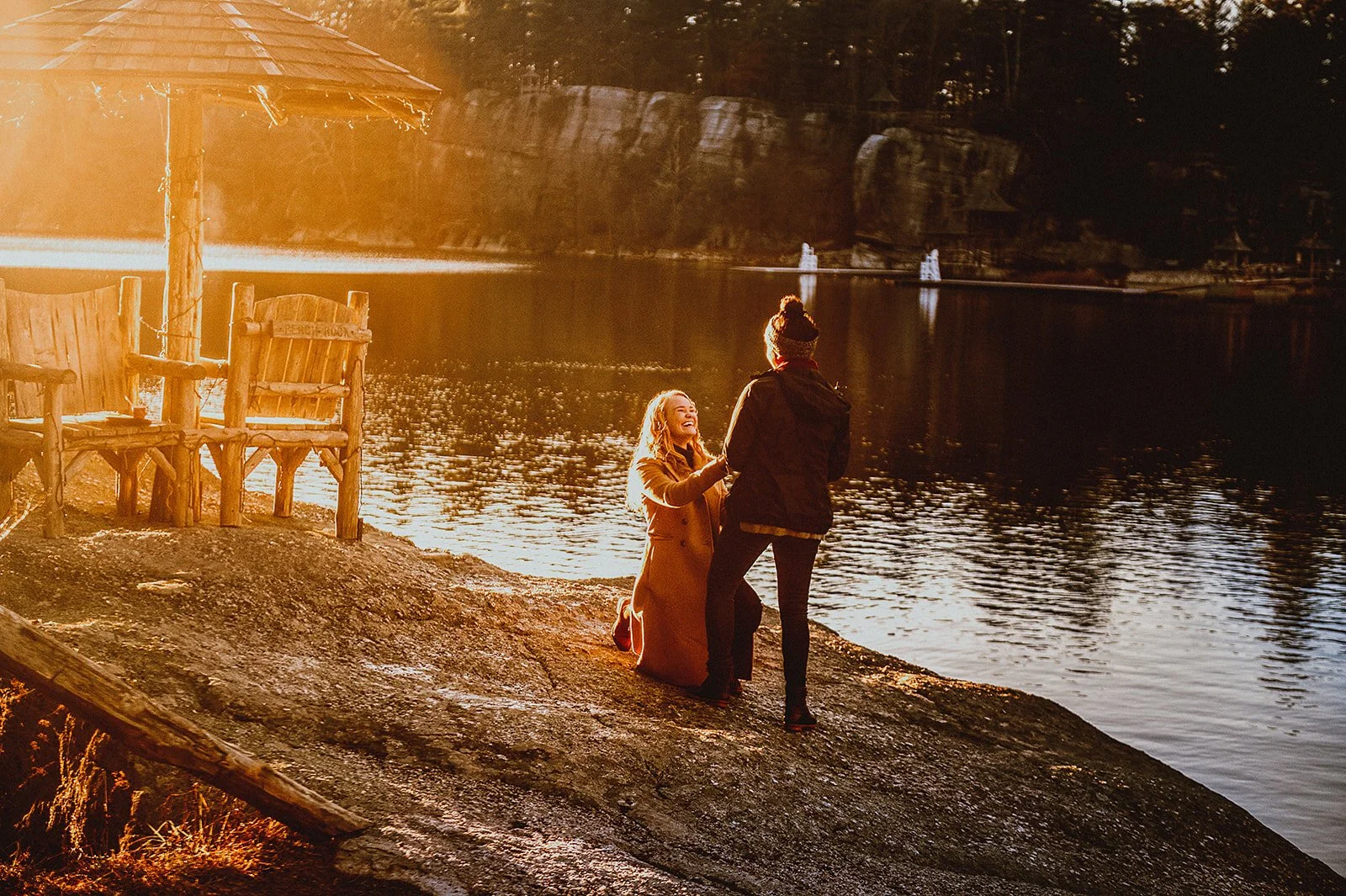 A man proposes to a woman on a rocky riverbank during sunset, with a wooden bench and a small shaded structure nearby, and water and trees in the background.