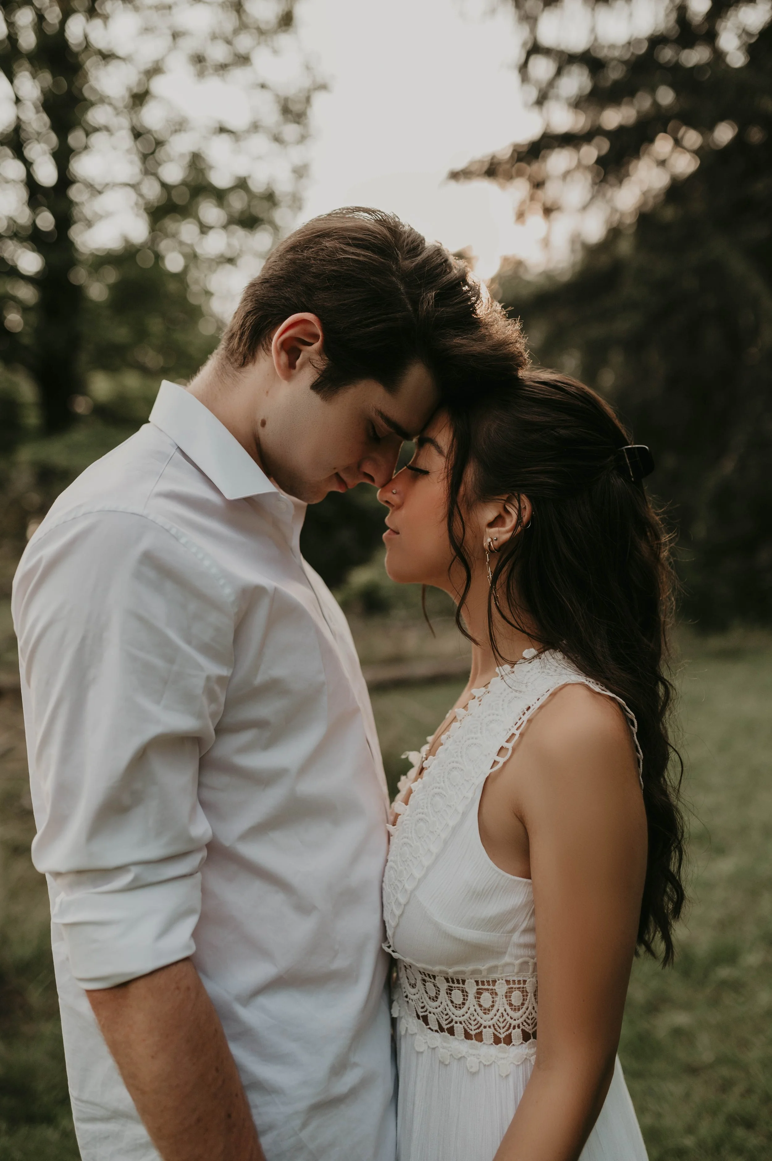 A young couple is standing outdoors, with their foreheads touching and eyes closed, in a tender moment. The man has brown hair and wears a white shirt, while the woman has long dark hair and wears a sleeveless white dress with lace details. The backg
