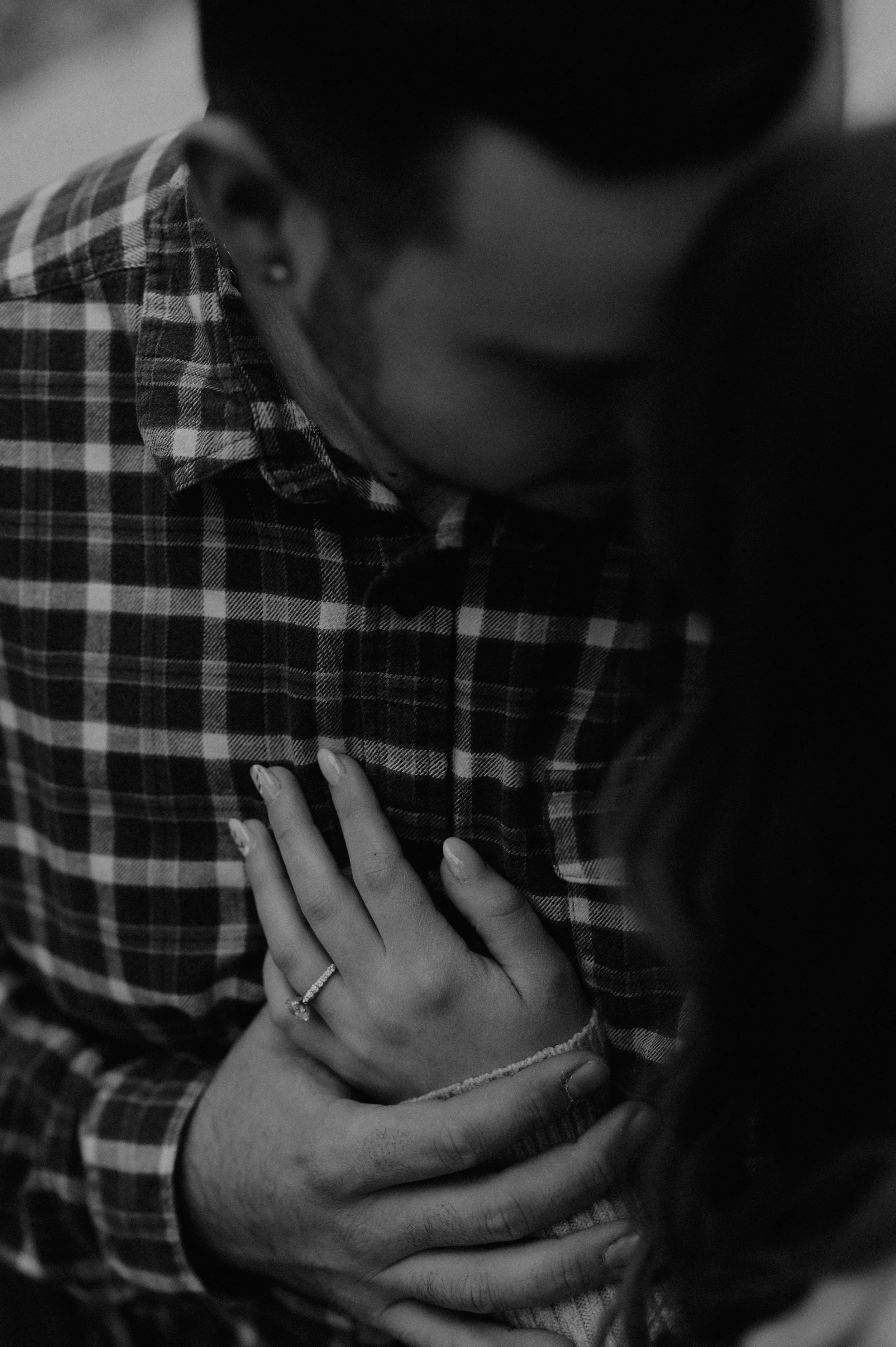 A black and white photo of a person with their arms crossed over their chest, wearing a plaid shirt and a wedding ring, with another person's hand resting on their shoulder.