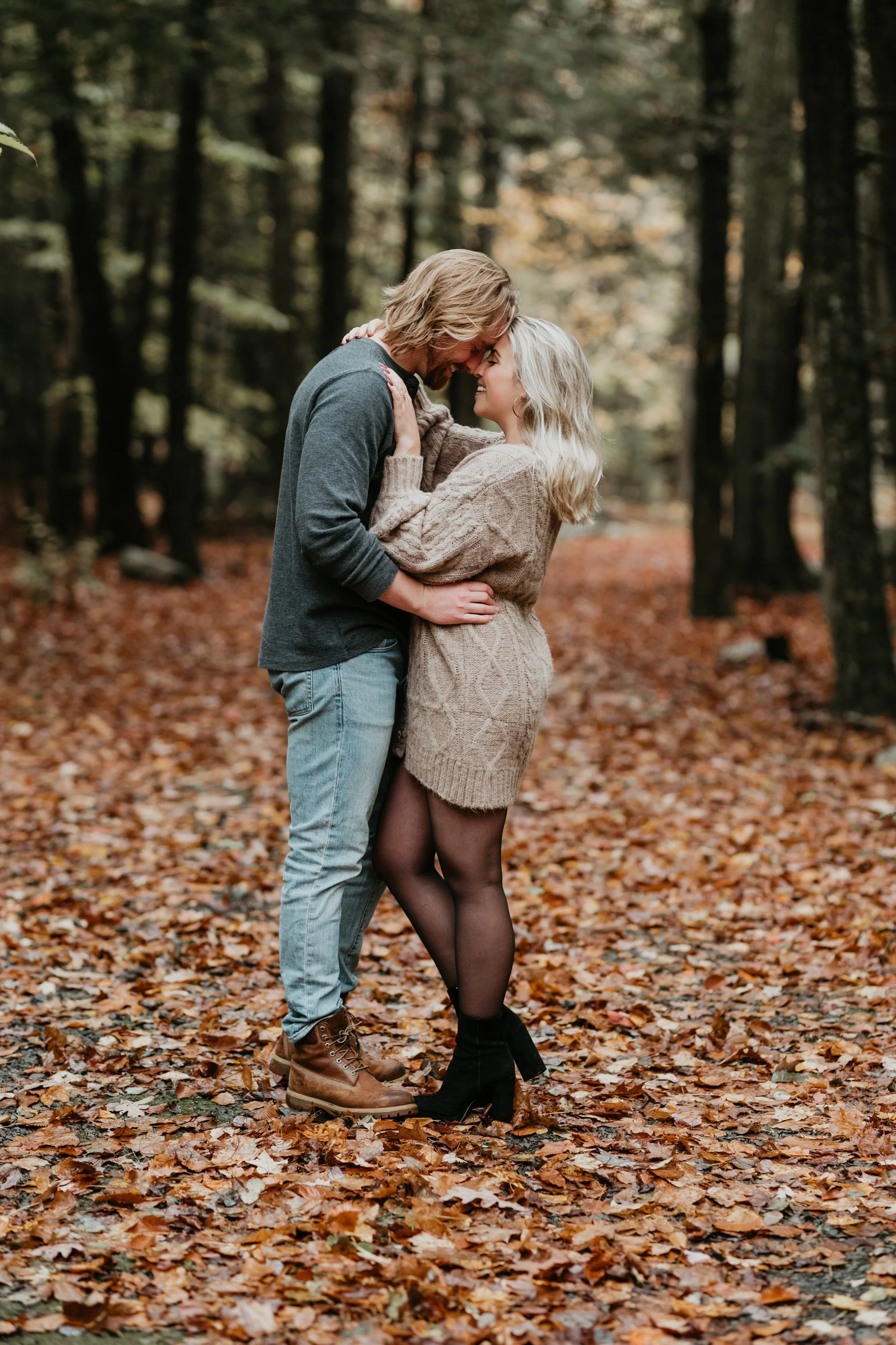 A couple embraces in a forest covered with autumn leaves, facing each other with foreheads touching.
