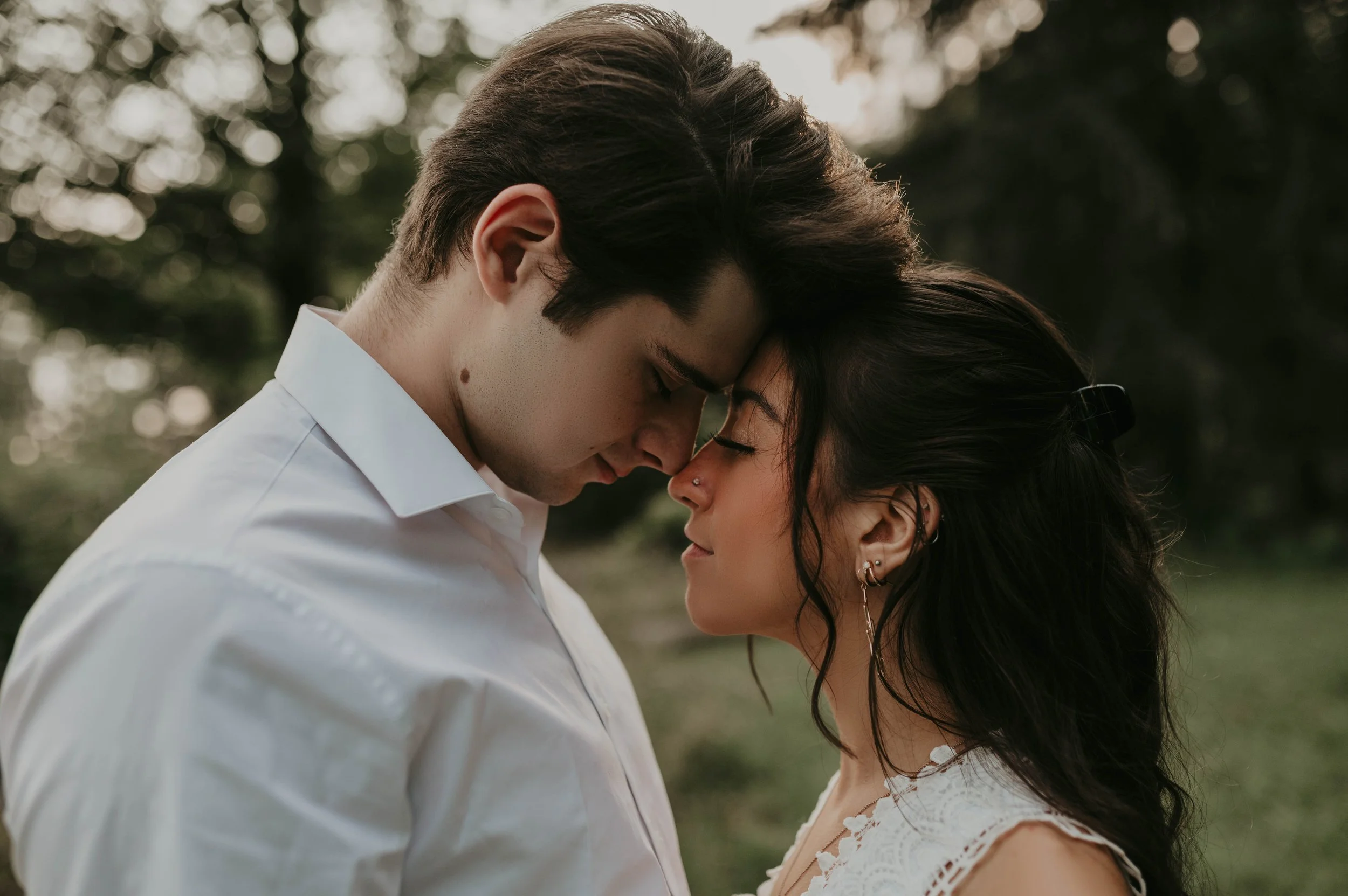 A young man and woman with their foreheads touching, eyes closed, outdoors in a tender embrace.