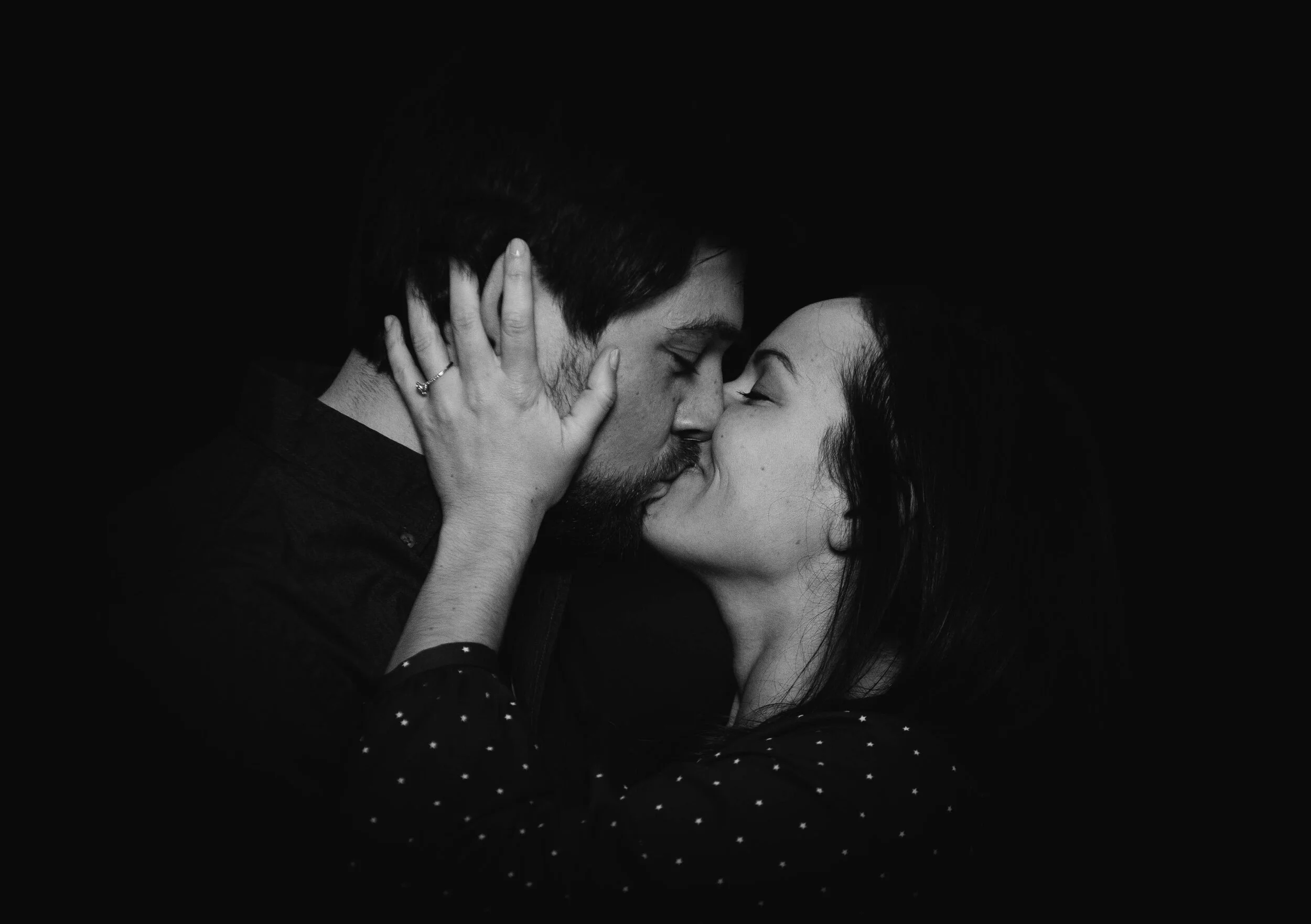 A black and white photo of a man and woman kissing with their eyes closed, woman holding man's face with one hand, against a dark background.