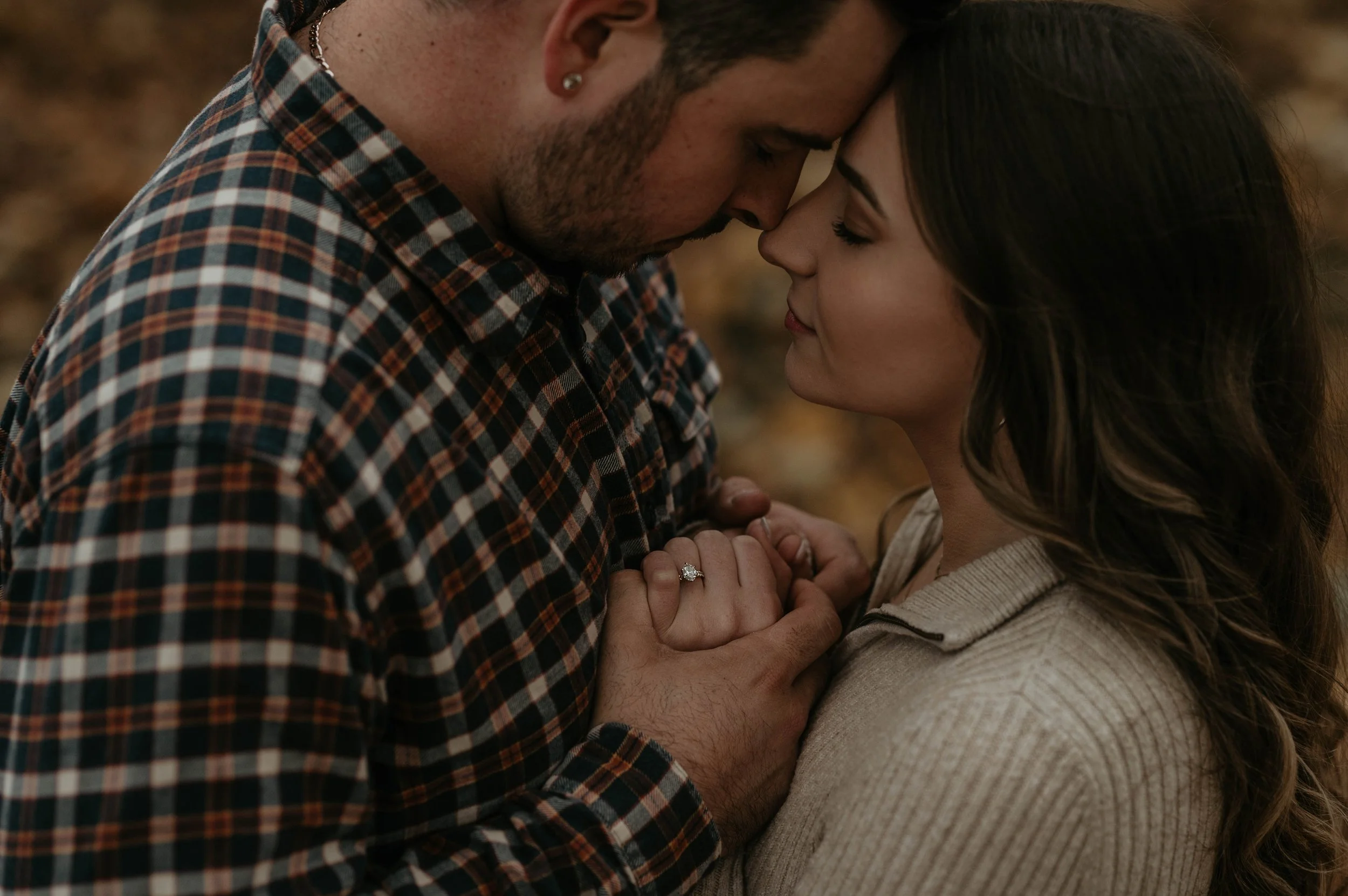 A couple is holding hands and touching foreheads, with the woman wearing an engagement ring, in an intimate embrace outdoors during autumn.